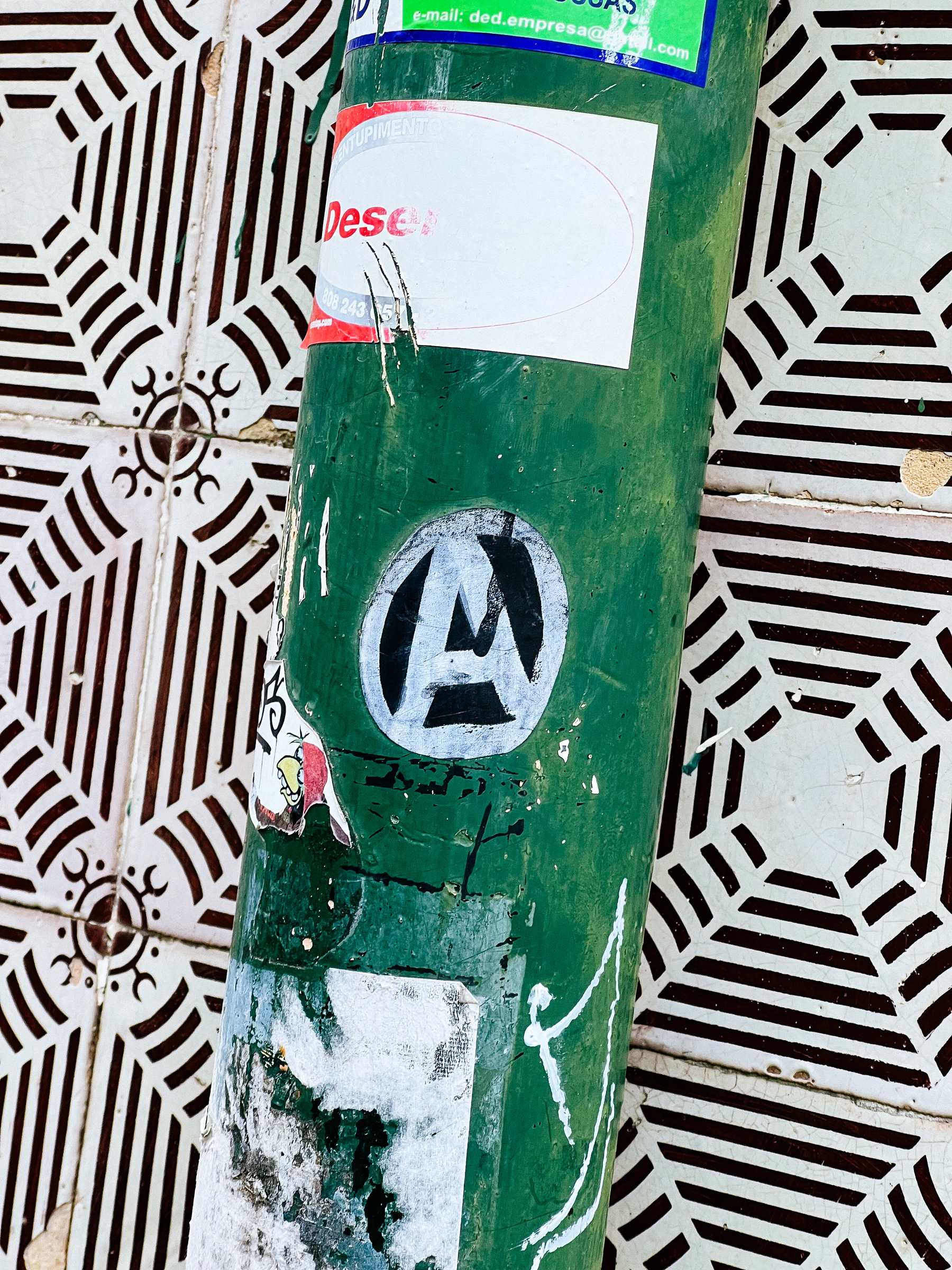 Anarchy. In a sticker. On a weathered green utility pole covered in layers of faded stickers, set against a backdrop of ornate, geometric-patterned tiles.