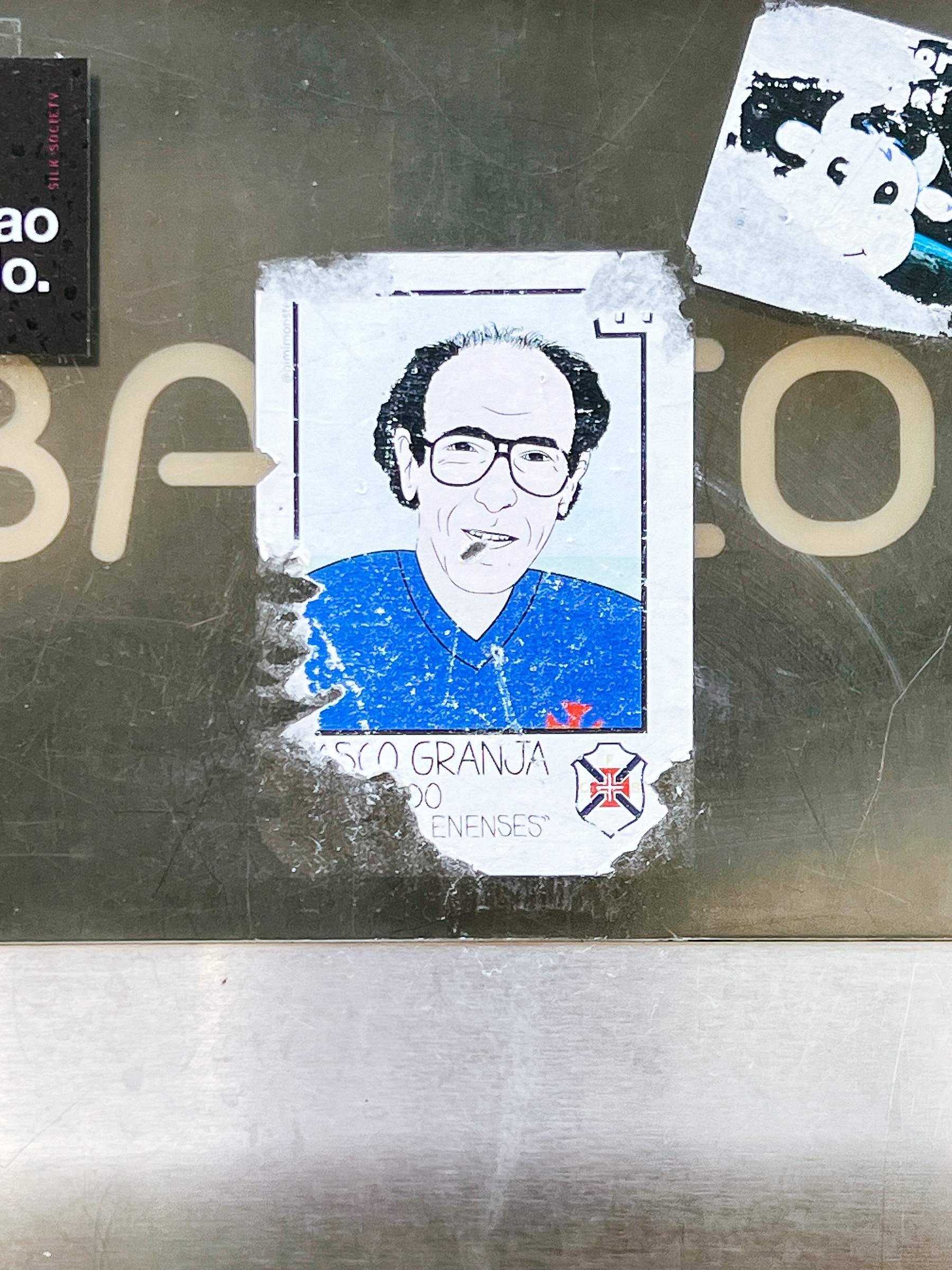 A sticker depicting a man&rsquo;s portrait is affixed to a metallic surface, surrounded by other partial stickers and graffiti.