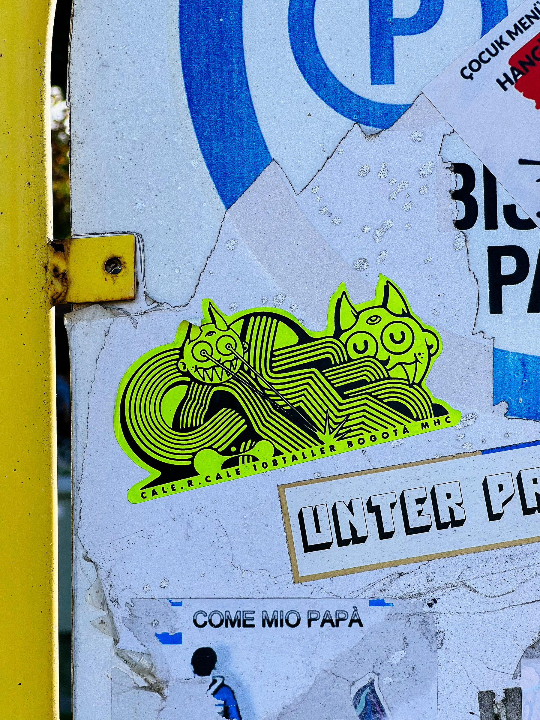 A neon green sticker depicting two stylized cats with large eyes is affixed to a weathered sign among other stickers and graffiti.