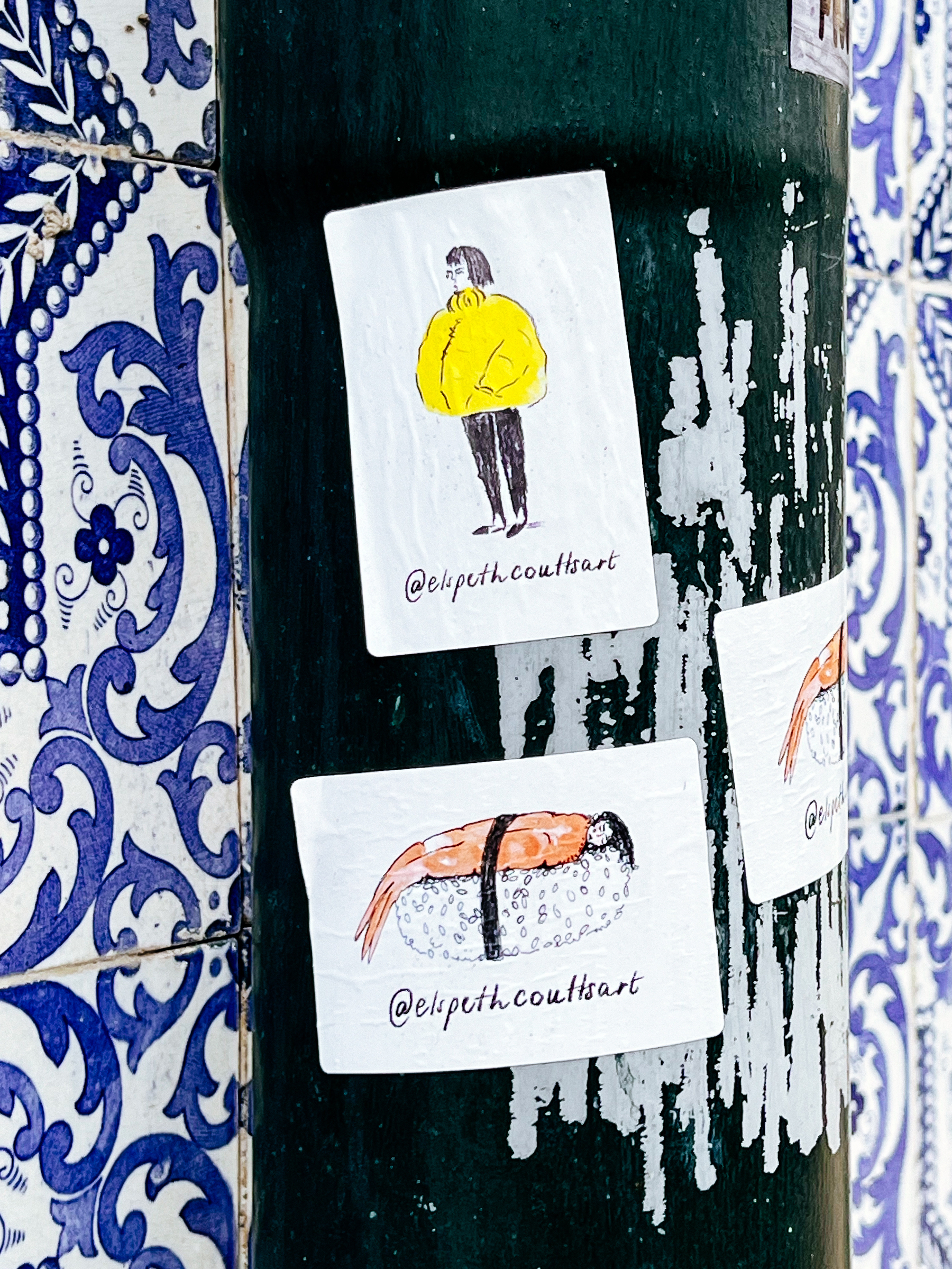 Stickers depicting a person in a yellow jacket and a person/shrimp sushi are affixed to a lamp post, with the artist’s handle displayed.