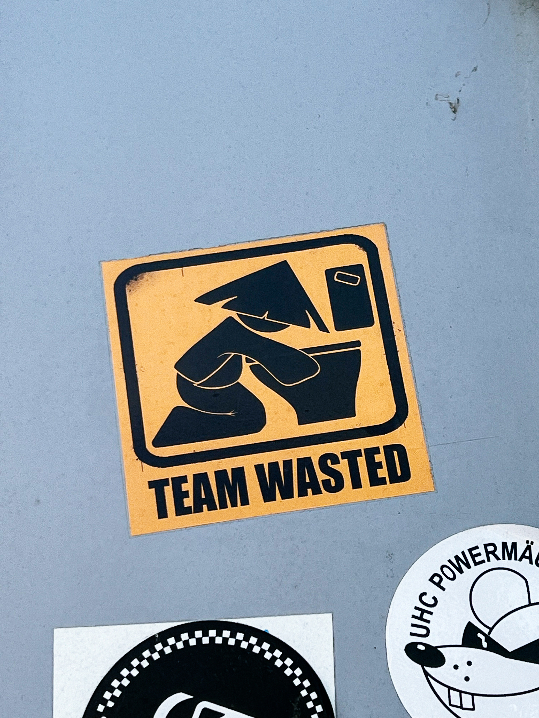 A sticker with a yellow background and a design of a person throwing up in a toilet bowl reads, &ldquo;TEAM WASTED,&rdquo; adhered to a grey surface alongside other stickers.