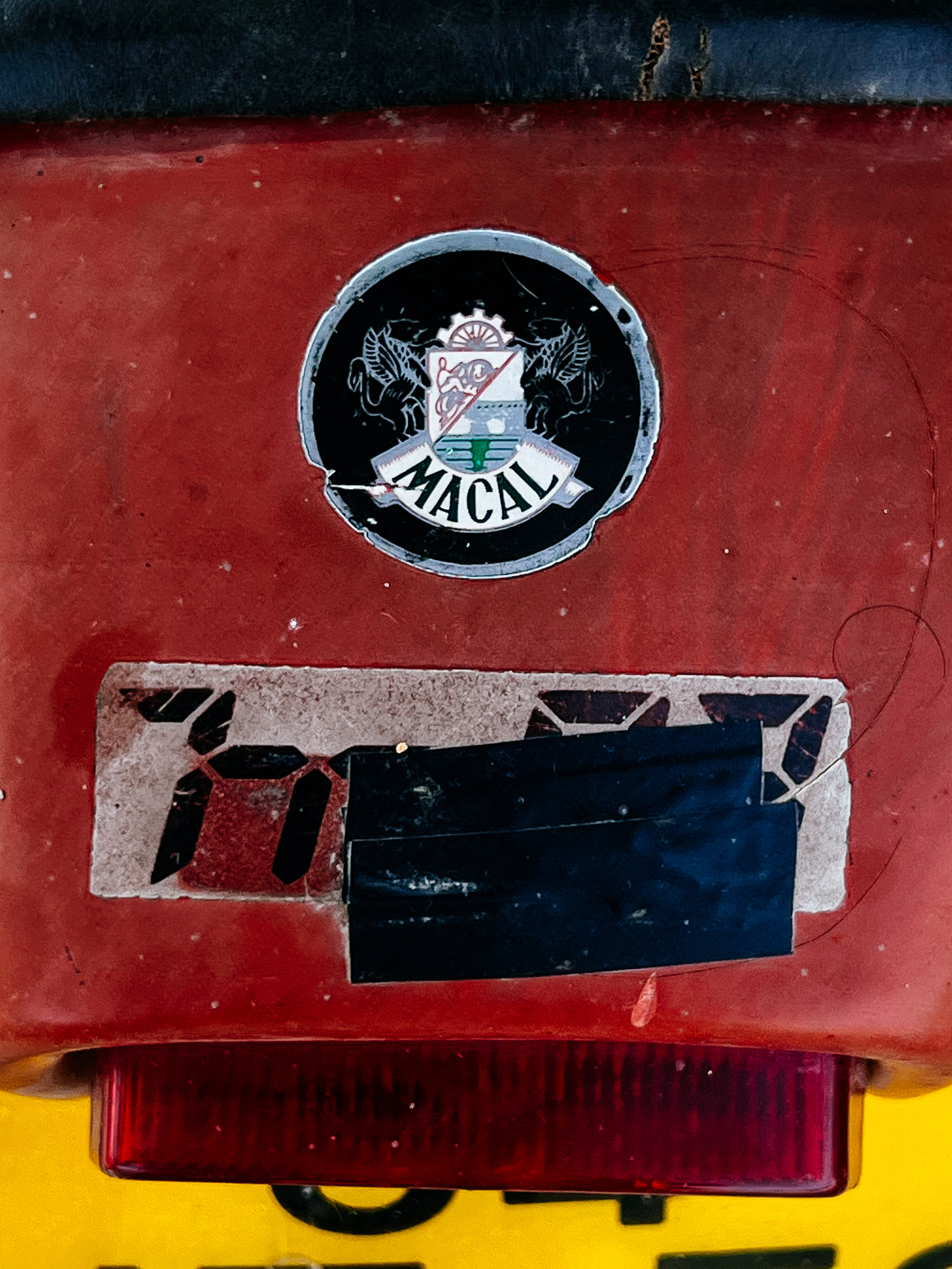A weathered emblem reads &ldquo;MACAL&rdquo; on a red surface above a taped, partially torn sticker, near a taillight on a yellow background.