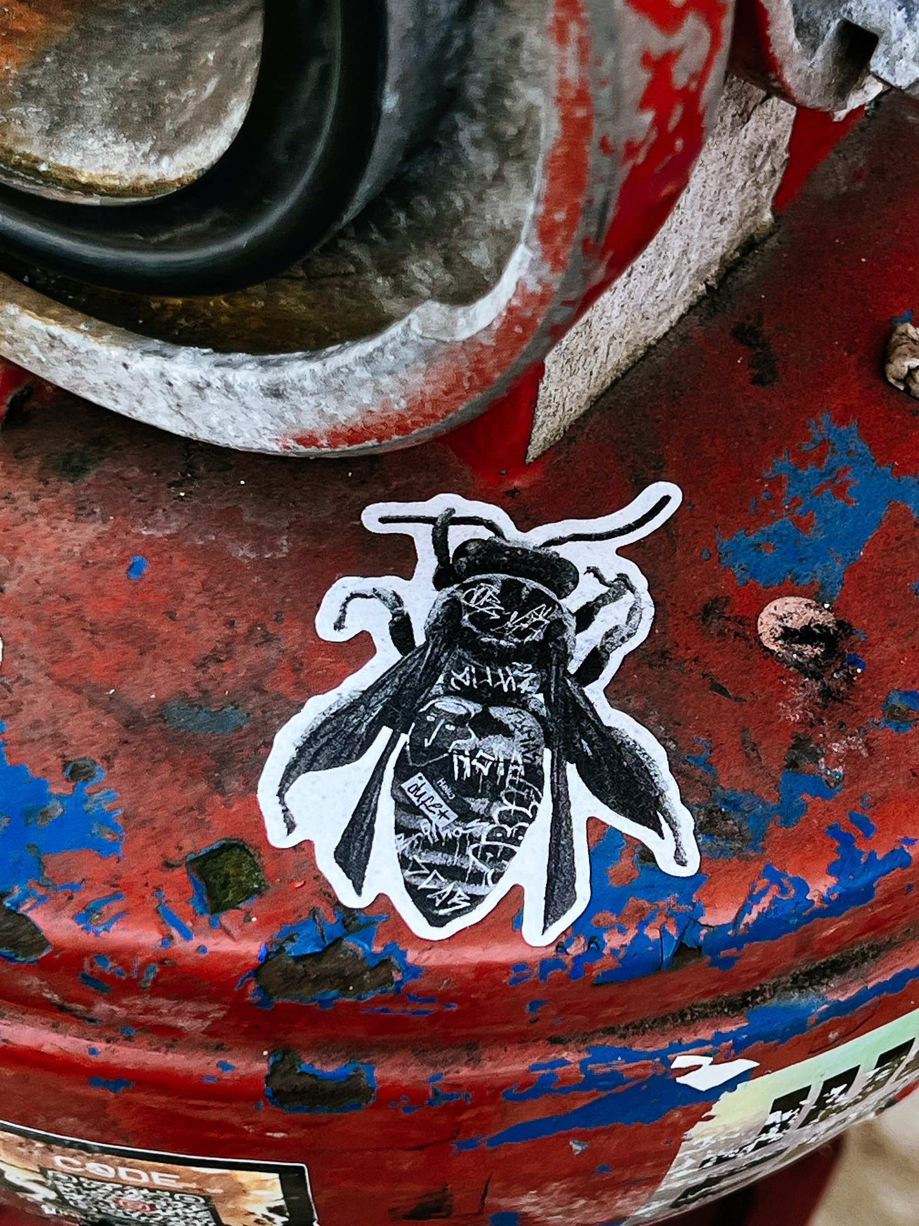 Sticker of a bee with intricate patterns adheres to a worn, red fire hydrant with peeling blue paint.