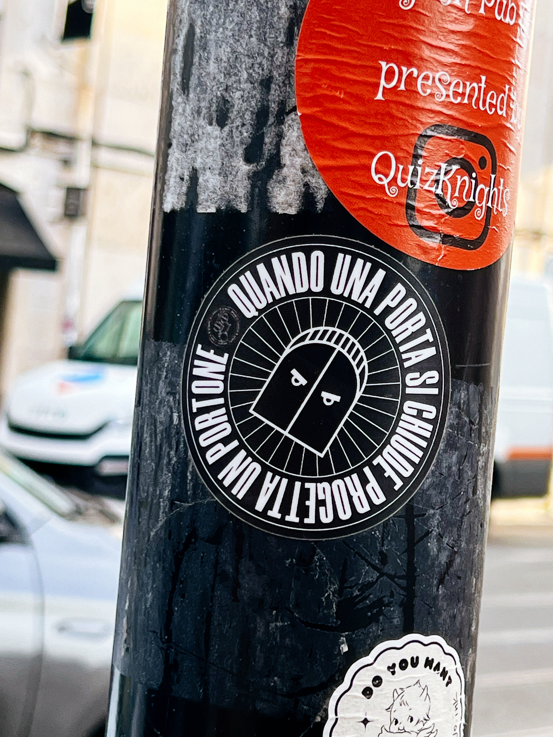 Stickers on a pole with one featuring text &ldquo;QUANDO UNA PORTA SI CHIUDE PROGETTA UN PORTONE&rdquo;, and a mean looking door.