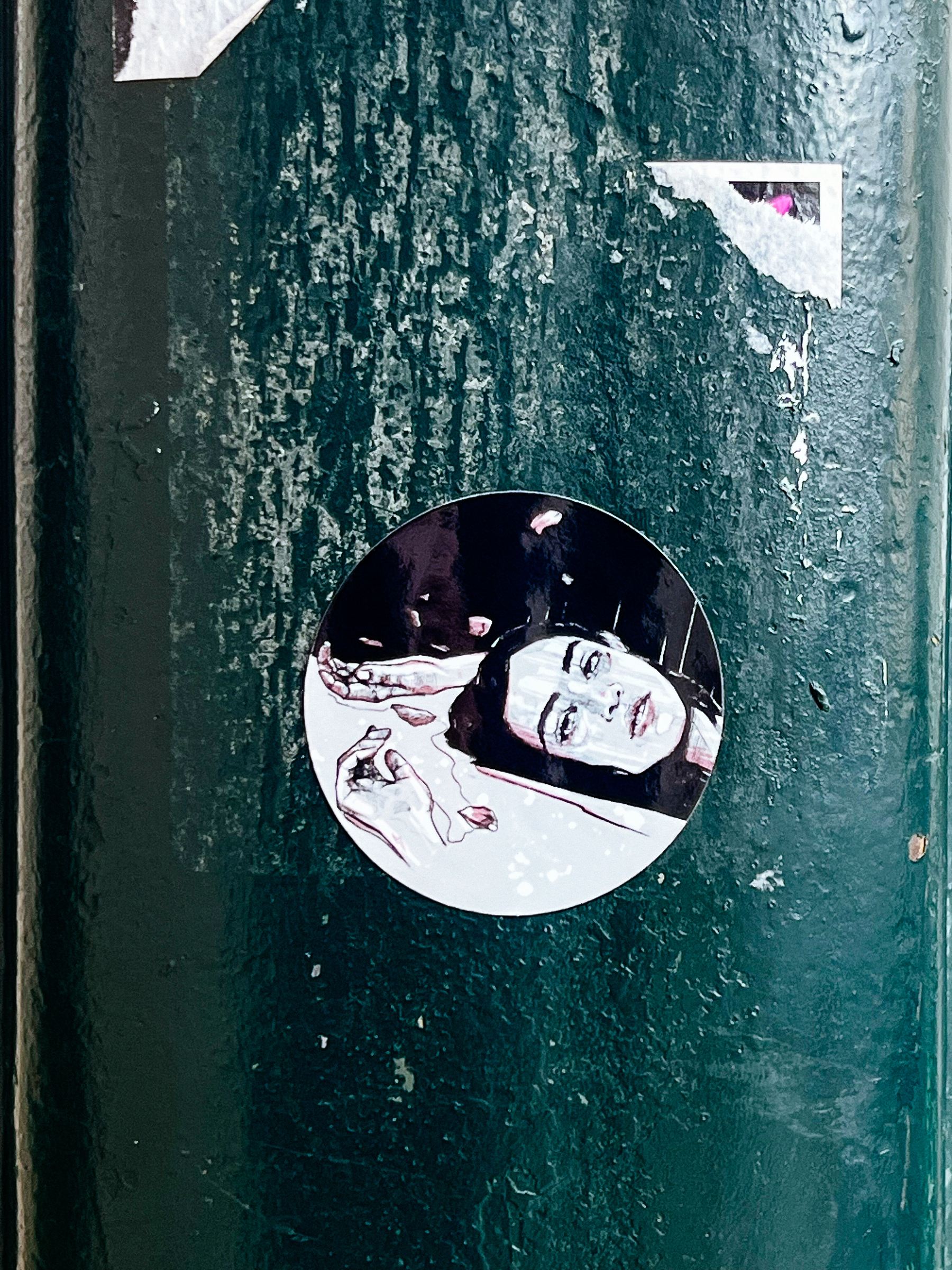 A sticker depicting a monochromatic illustration of a person&rsquo;s face and hand is adhered to a textured green surface, surrounded by remnants of other stickers.