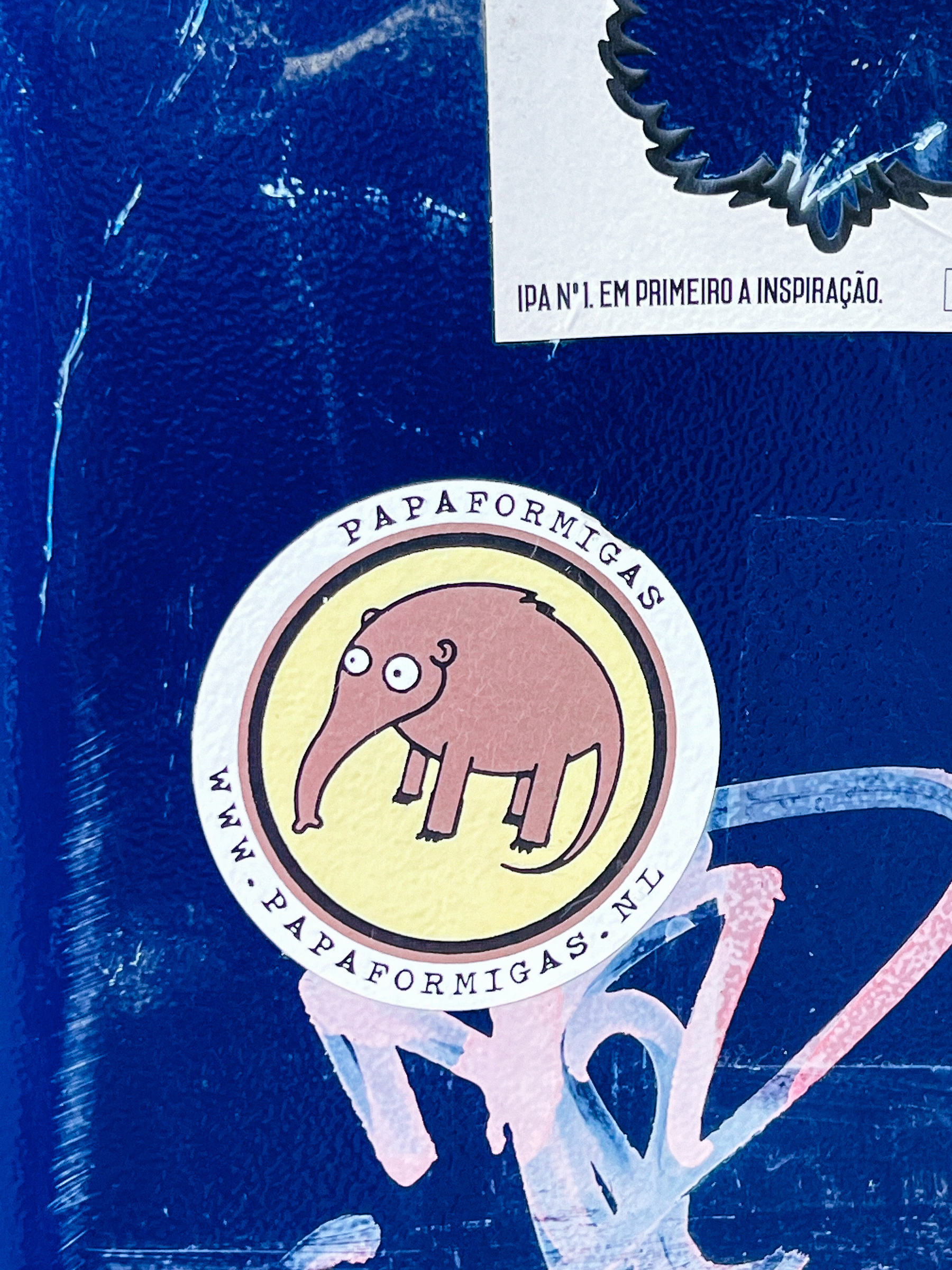 A sticker of a stylized anteater with &ldquo;PAPAFORMIGAS&rdquo; and &ldquo;WWW.PAPAFORMIGAS.NL&rdquo; is affixed to a blue surface with other graffiti.