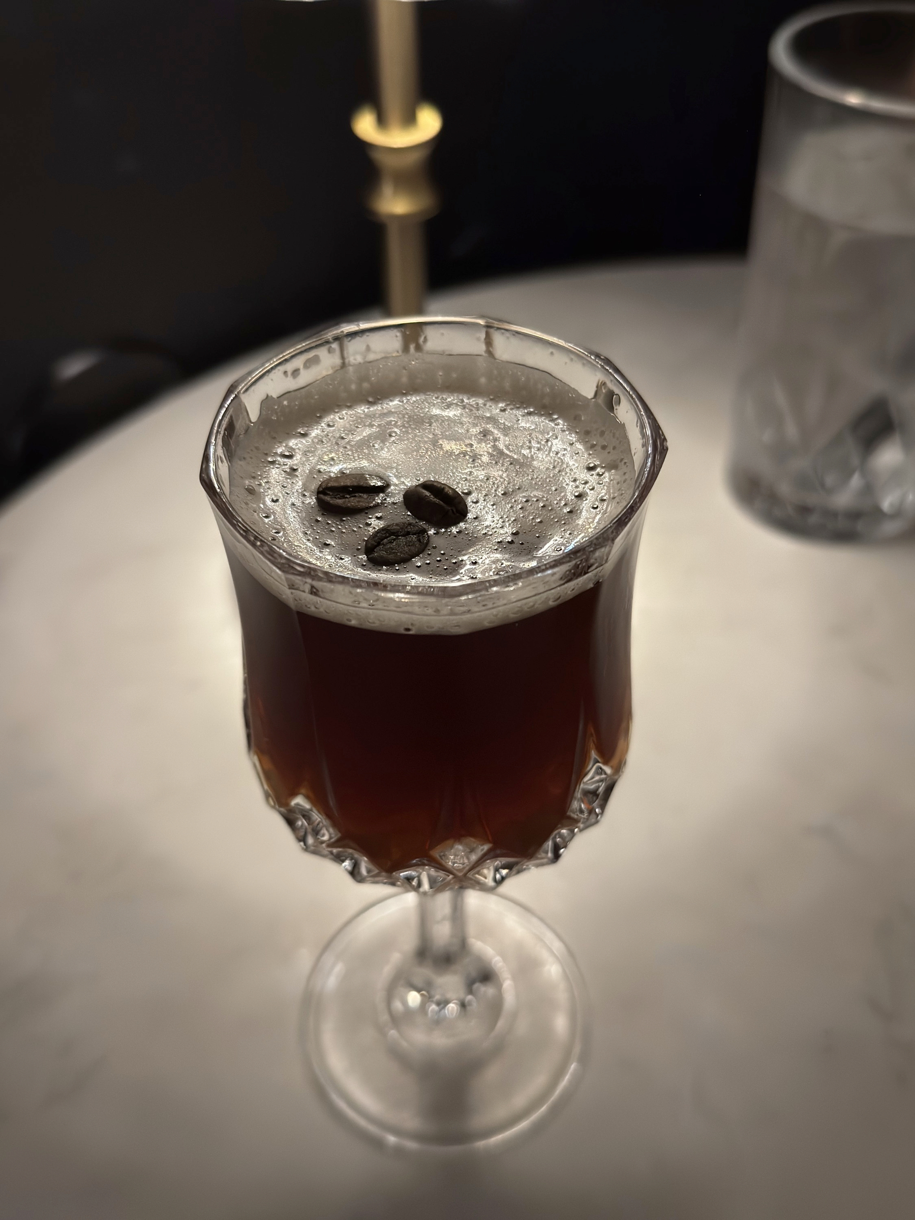 A dark coffee cocktail is served in a faceted crystal goblet, topped with a delicate layer of foam and three glossy coffee beans, the rich brown liquid catching warm lamplight and looking smooth, aromatic, and inviting.
