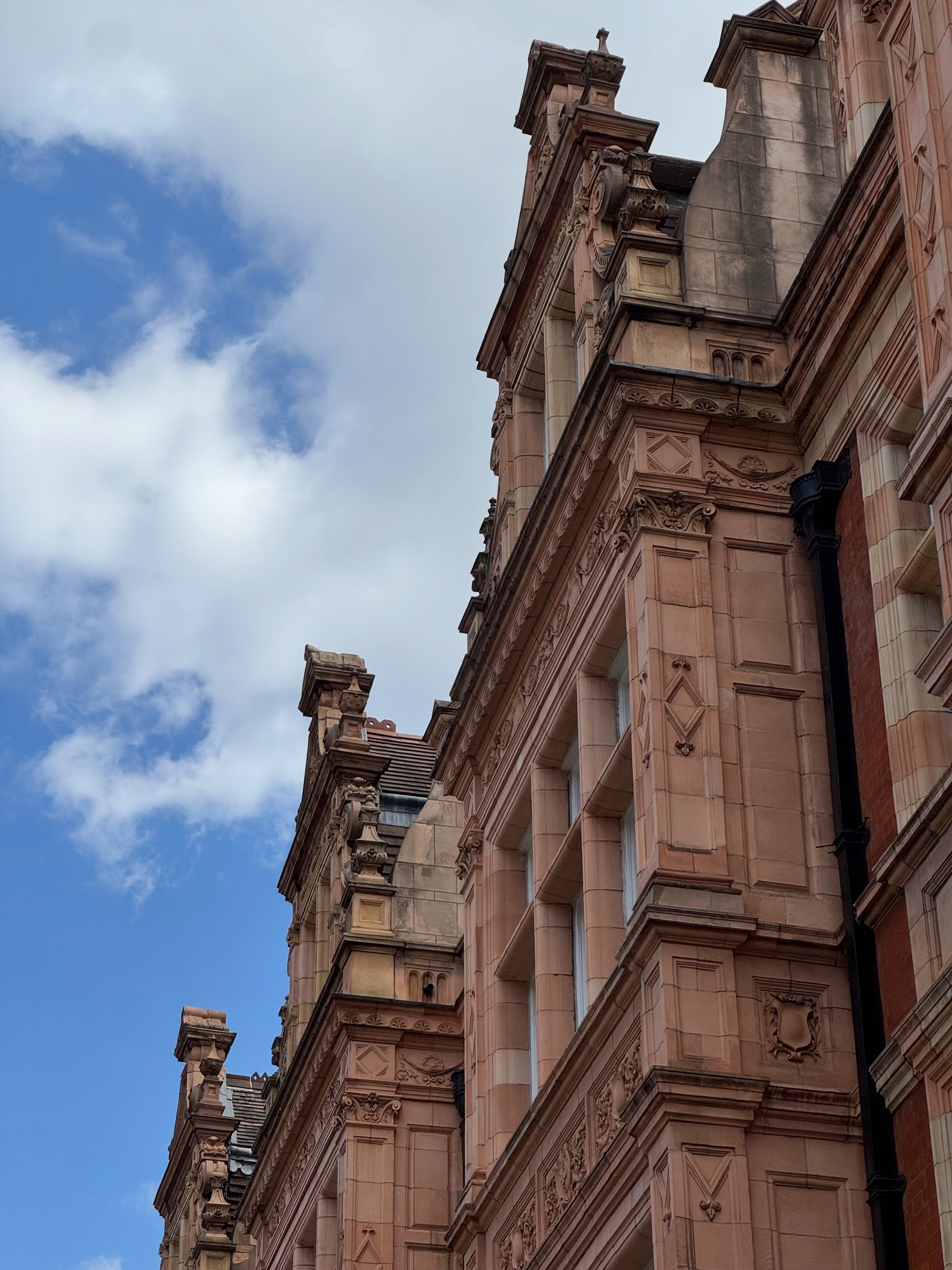 Detailed stone facade of a grand Edwardian building, viewed from below to emphasize carved columns, cornices, and sculptural details rising toward a cloudy blue sky.