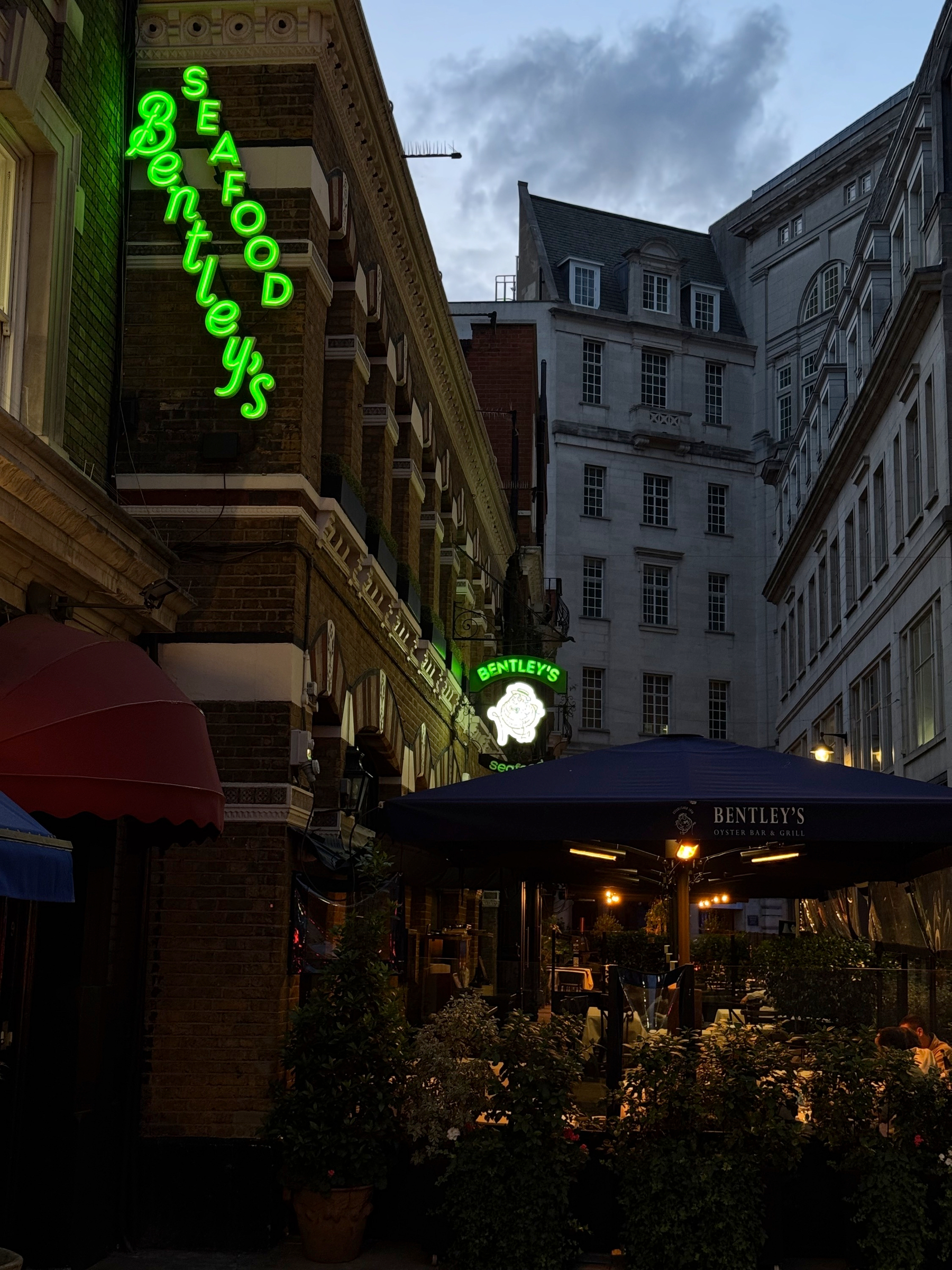 The restaurant exterior at dusk, with glowing green neon signage and warmly lit outdoor seating tucked into a narrow London street, creating an inviting contrast against the darkening sky.