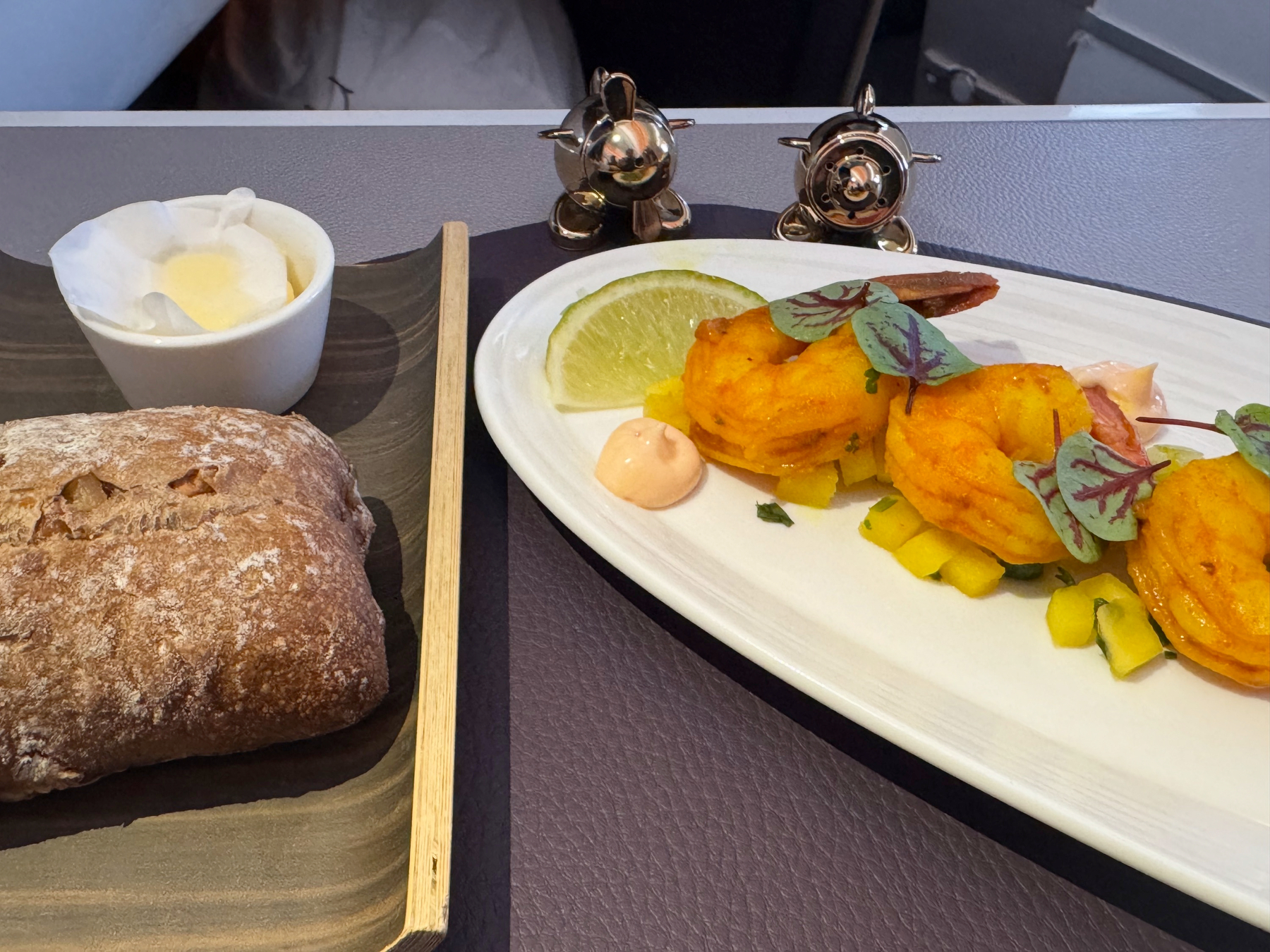 An elegant starter featuring golden shrimp arranged with bright yellow garnish, microgreens, a lime wedge, and a smooth pink sauce, served alongside rustic bread and butter.