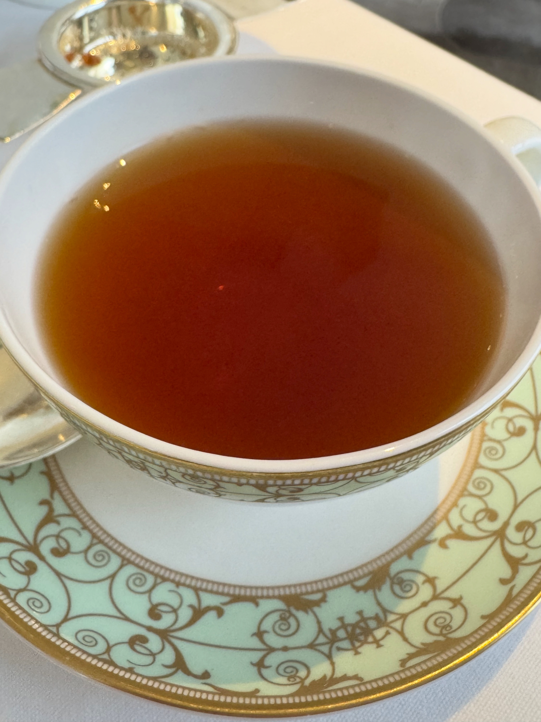 Amber-colored tea fills a delicate patterned teacup and saucer, the surface clear and glowing under warm light. The fine porcelain and gold detailing emphasize the calm, unhurried ritual of tea service.