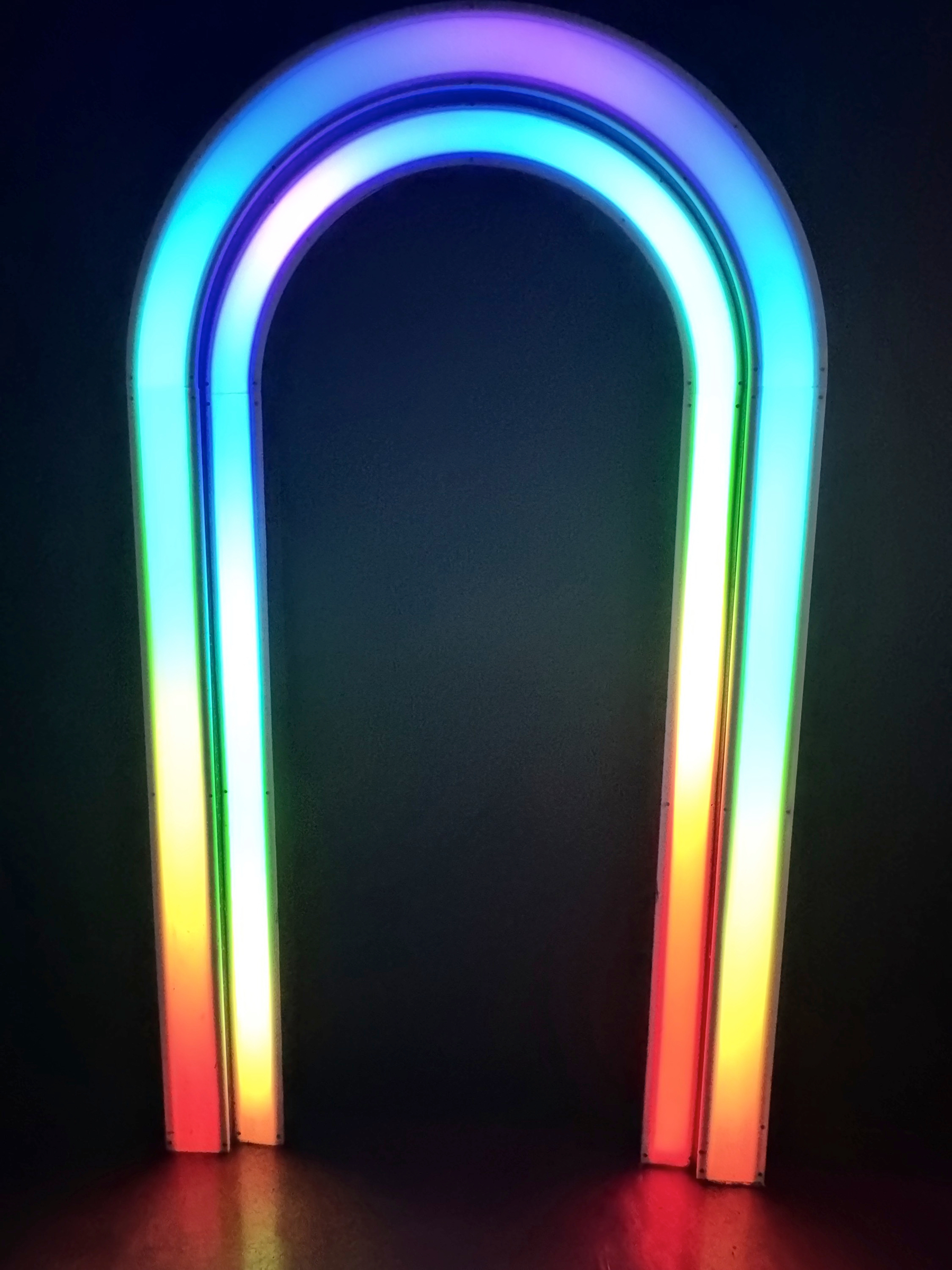 A glowing, inverted U-shaped neon arch stands against a dark wall, its smooth bands transitioning from violet and blue at the top to green, yellow, and red near the base, creating a luminous rainbow halo that reflects softly on the floor.