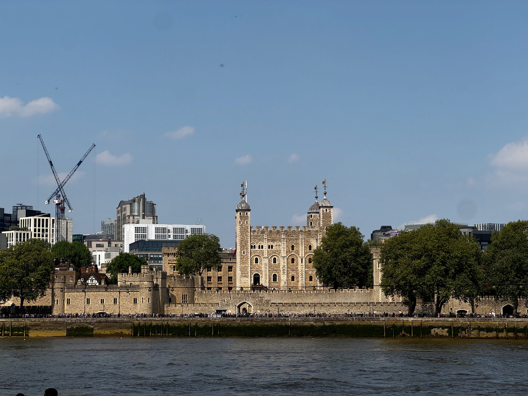 The White Tower and surrounding stone walls rise behind the River Thames under a clear blue sky, with leafy trees lining the ramparts and modern London buildings and cranes visible in the distance, creating a striking contrast between medieval architecture and the contemporary skyline.