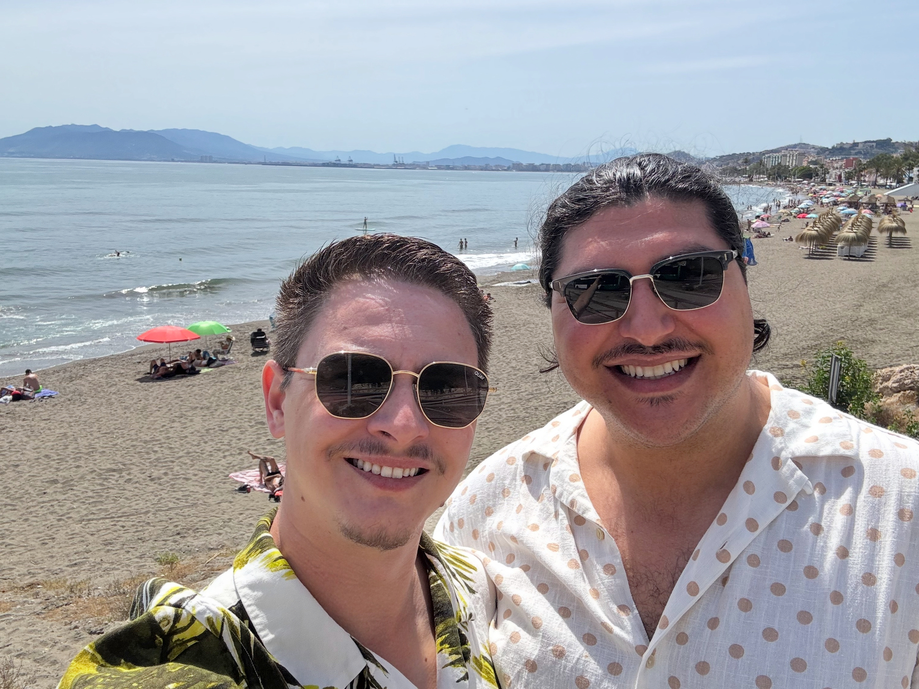 Hugo and I smiling for a close selfie on a sandy beach, with calm Mediterranean water, distant mountains, and scattered umbrellas and sunbathers stretching along the shore behind us.