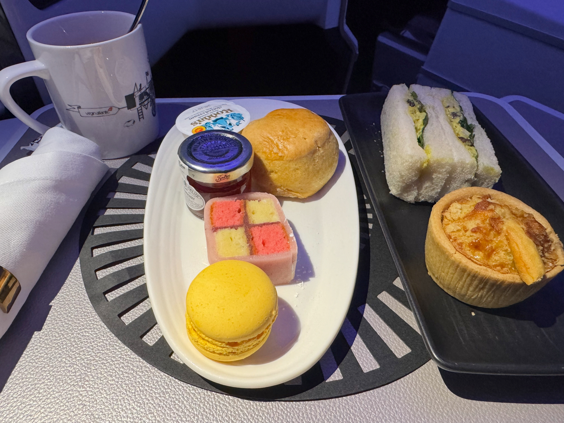 An afternoon tea spread with finger sandwiches, a warm scone with jam and clotted cream, a lemon macaron, a Battenberg-style cake, and a small tart, served with tea.
