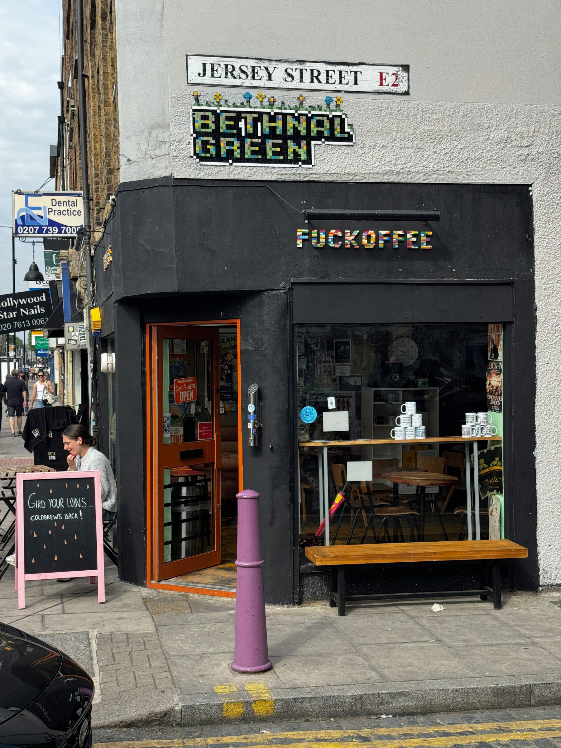 Corner café on Jersey Street with a black-painted façade, a pixel-style sign reading “F*ckoffee,” and a mosaic “Bethnal Green” sign above, with a bench, window display of mugs, and pedestrians passing by on the pavement.