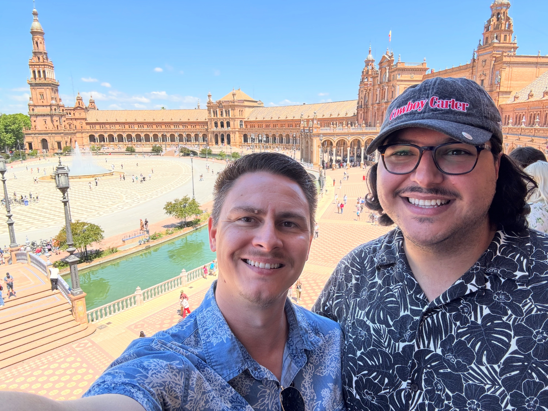Hugo and I smiling at the camera from an elevated viewpoint overlooking Plaza de España, with its sweeping brick buildings, tiled bridges, canal, and central fountain filled with visitors.