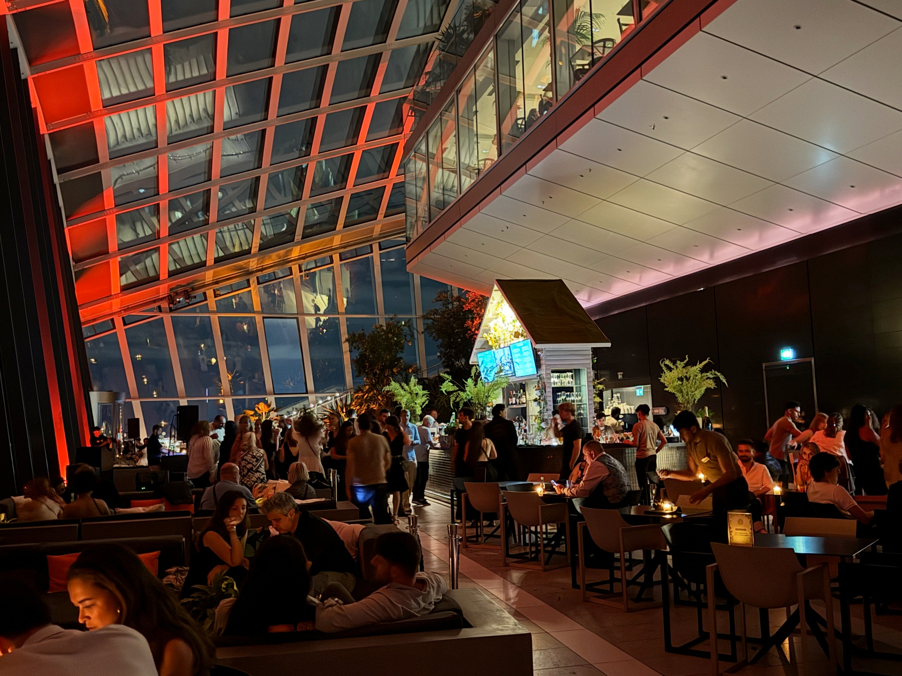A busy indoor garden bar with a slanted glass ceiling glowing red and orange, where groups sit at tables and lounge seating while others stand near the bar, surrounded by plants and city lights beyond the windows.
