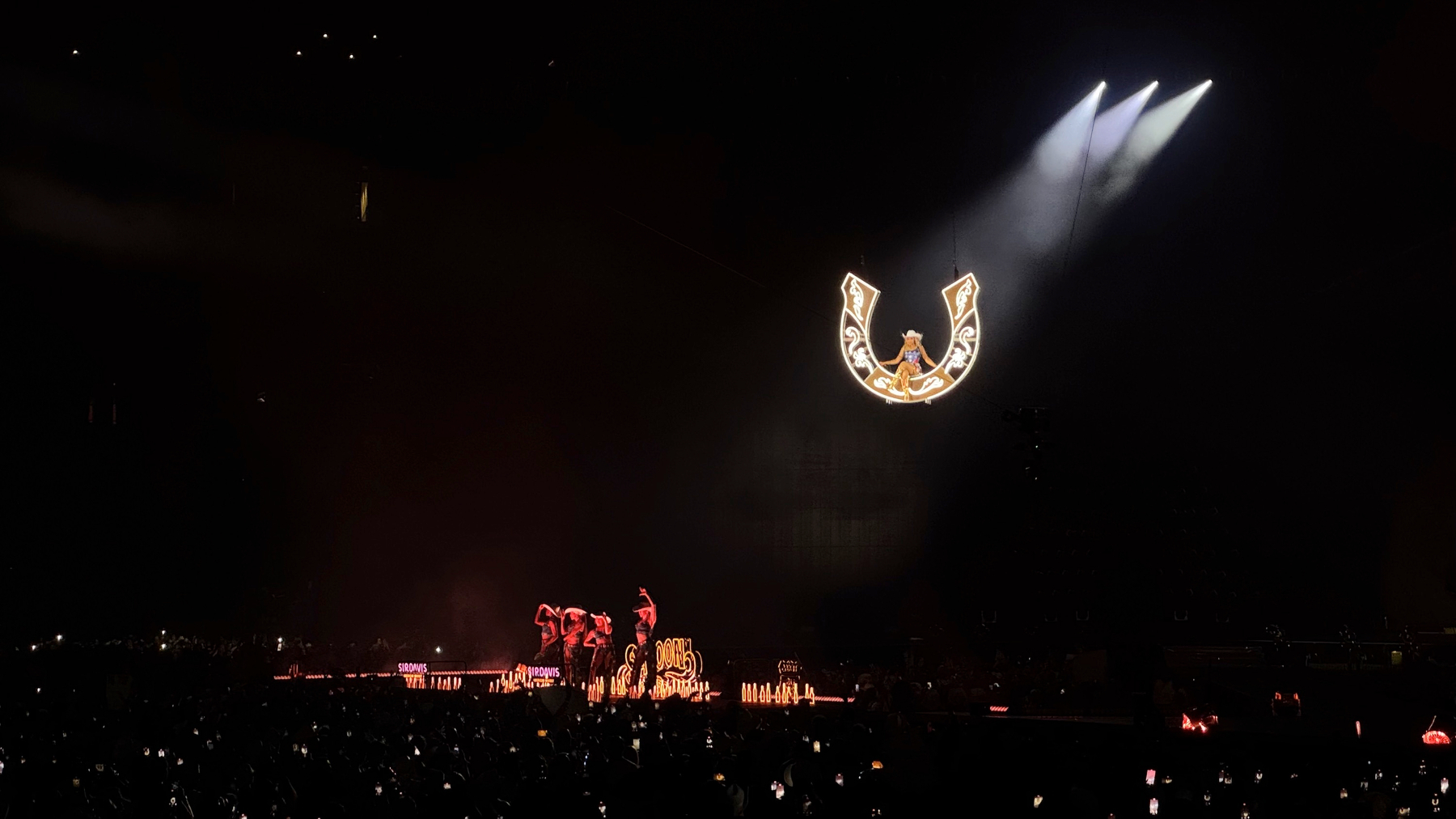 Beyoncé appears elevated high above the stage, seated within a glowing illuminated horseshoe suspended in darkness. Spotlights cut through the blackened stadium, isolating her as the focal point while the crowd below watches, their phone screens twinkling like stars.