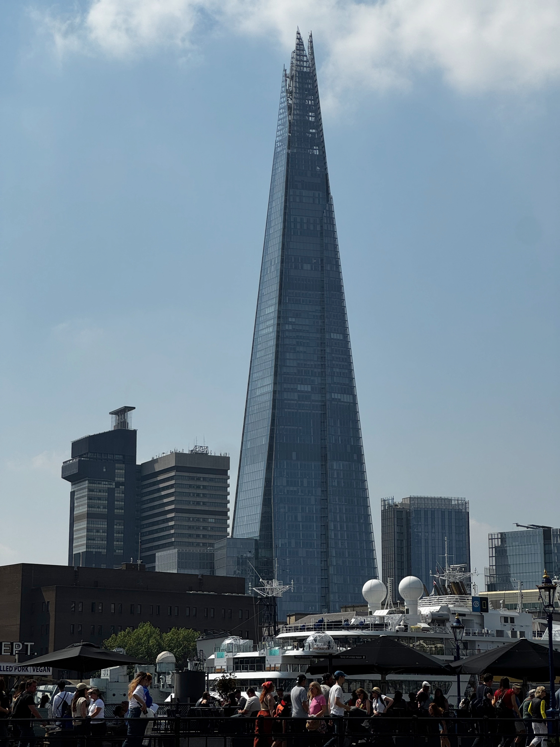 The Shard rising sharply above surrounding buildings, its glass surface reflecting daylight, with crowds gathered along the riverfront below and boats and rooftops filling the lower frame.