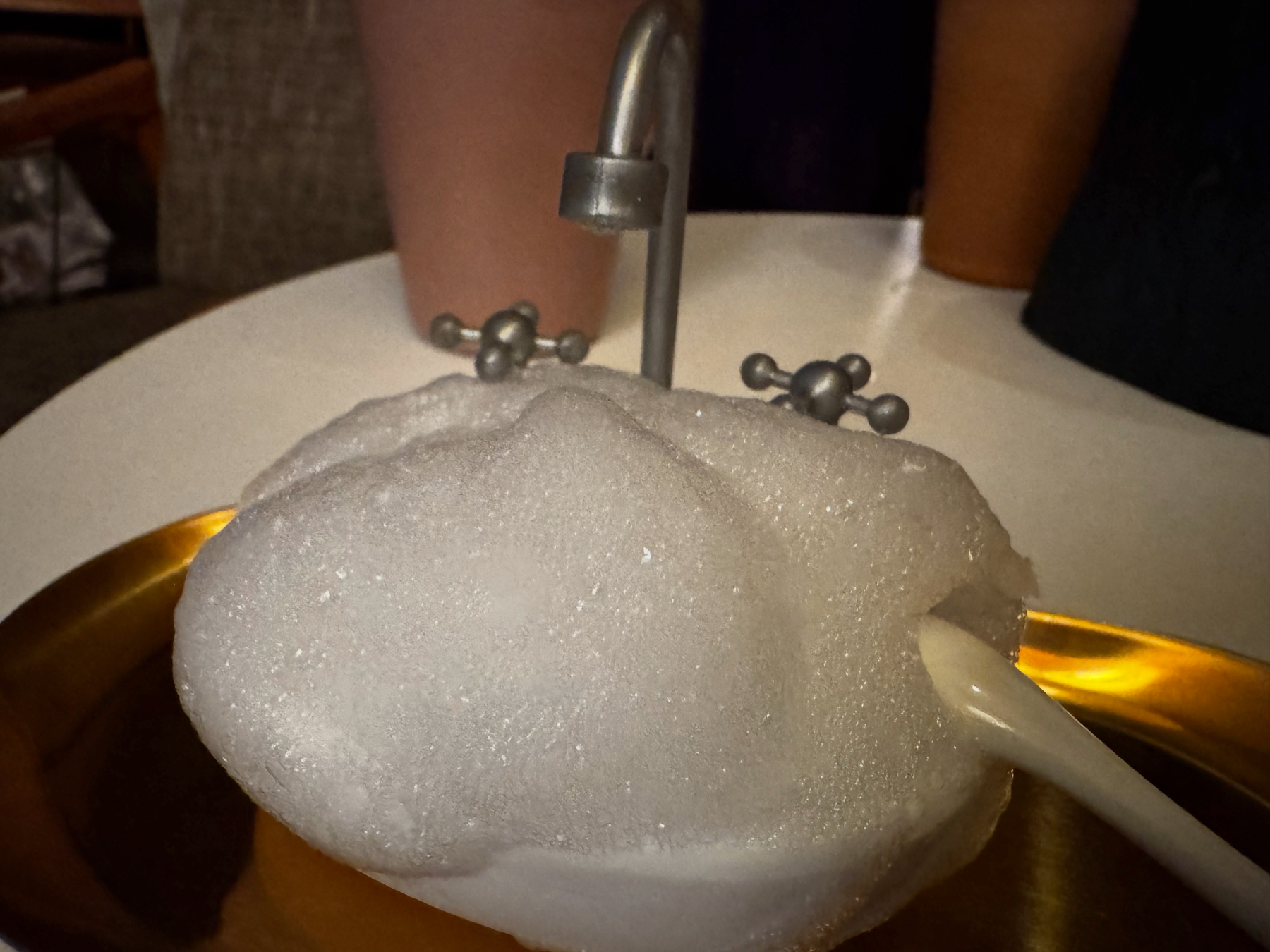 A glass topped with a thick, glossy cap of white foam, pierced by a tiny metal tap and spoon, creating a whimsical, interactive drink that looks rich, creamy, and meticulously constructed.