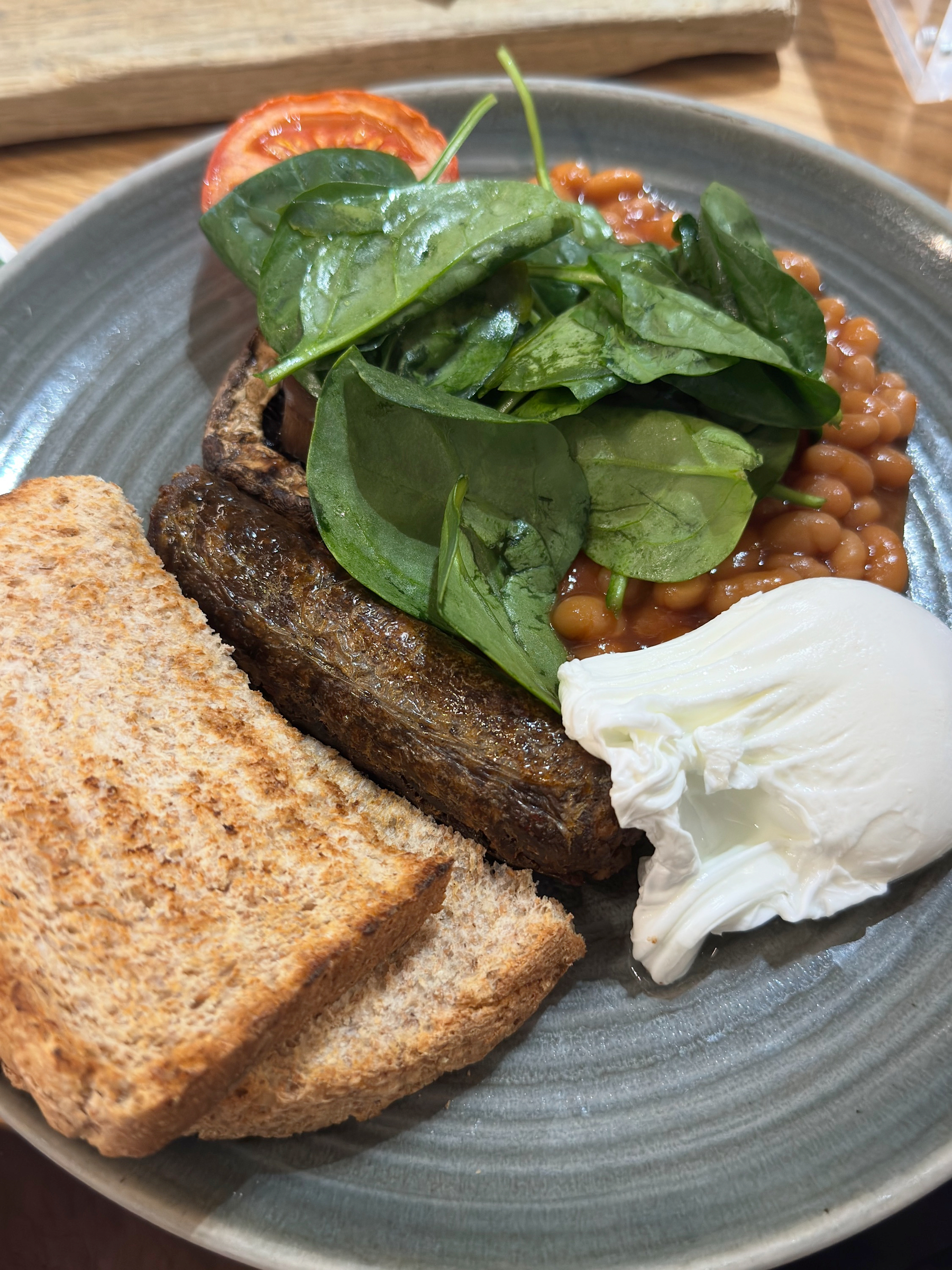 A hearty breakfast plate with toasted brown bread, a thick grilled mushroom, baked beans, wilted spinach, a slice of tomato, and a softly poached egg with a glossy white.