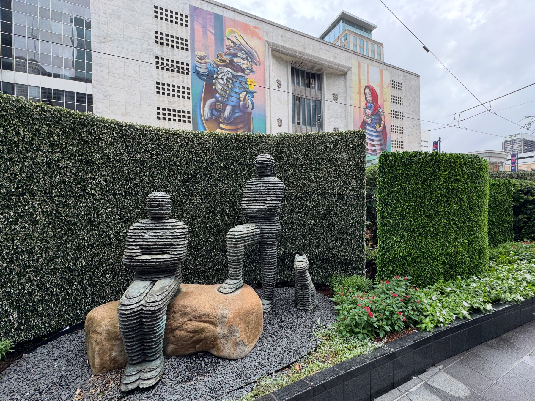 Abstract layered sculptures of seated and standing human figures arranged in a landscaped garden, with manicured hedges and a large colorful mural on a civic building visible in the background.