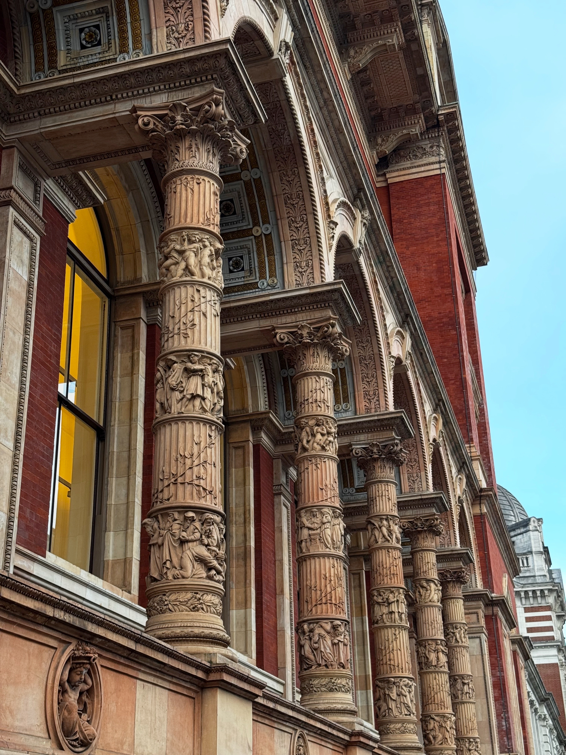 Row of richly carved stone columns on a red-brick façade, each column wrapped with detailed relief figures and decorative patterns, creating a dense, ornate rhythm along the museum exterior.