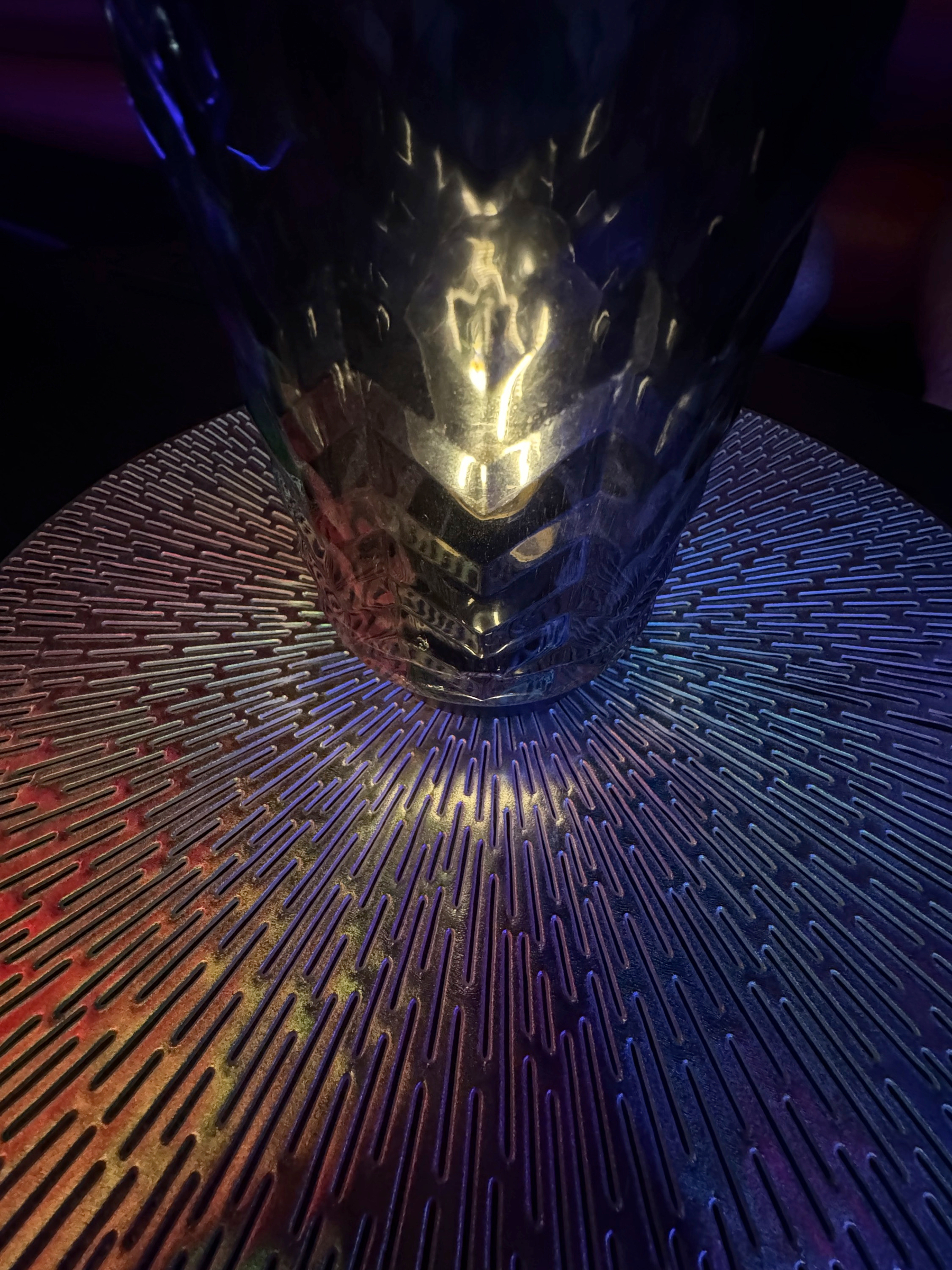 A close-up of a tall, textured glass catches dramatic blue and amber light, the rippled surface refracting a warm glow onto a patterned tabletop, creating a moody, abstract play of reflections and color.