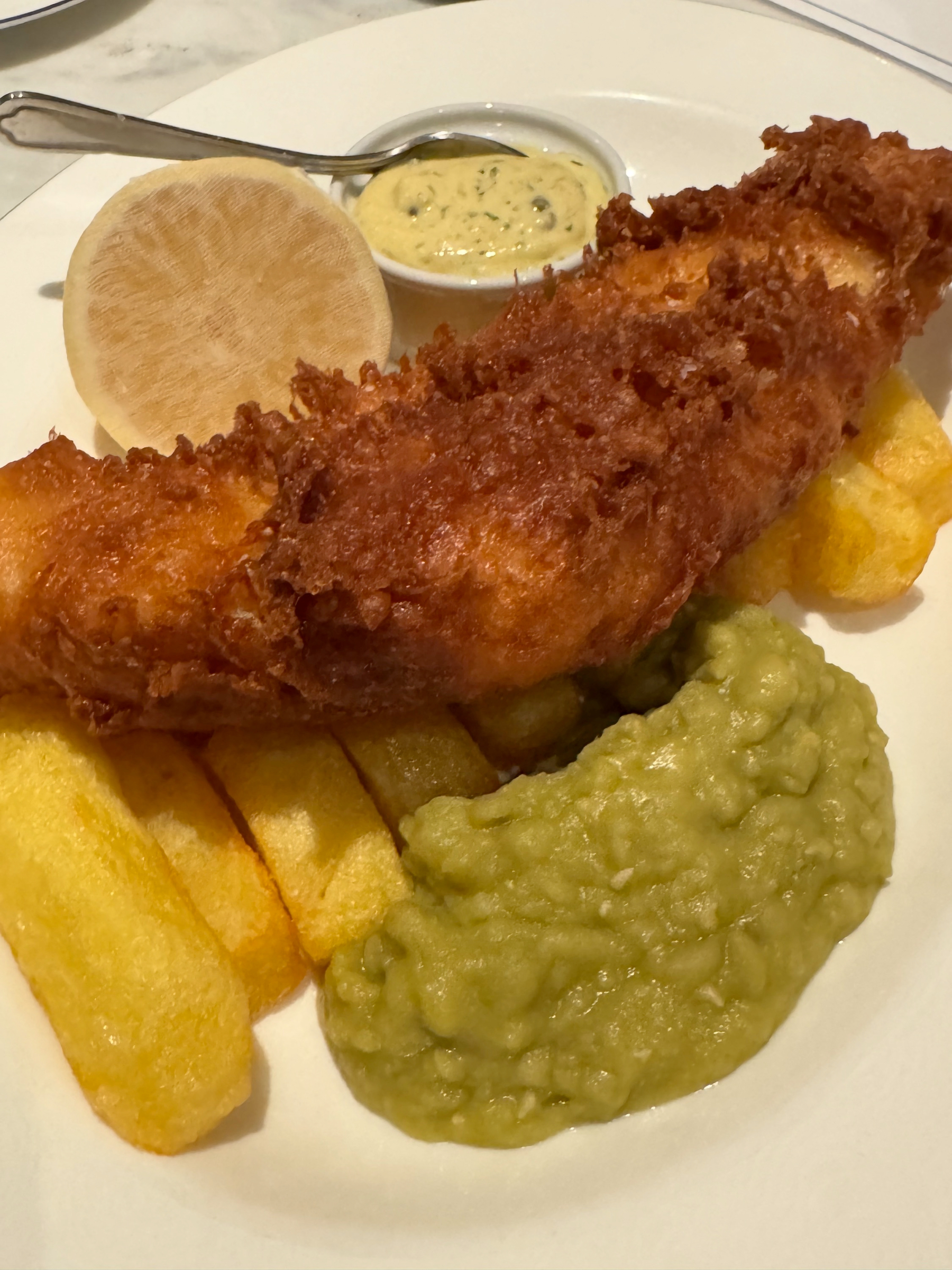 Golden battered fish resting over thick-cut chips, the crust deeply crisp and textured, served with mushy peas and a pot of tartar sauce for a classic, comforting plate.