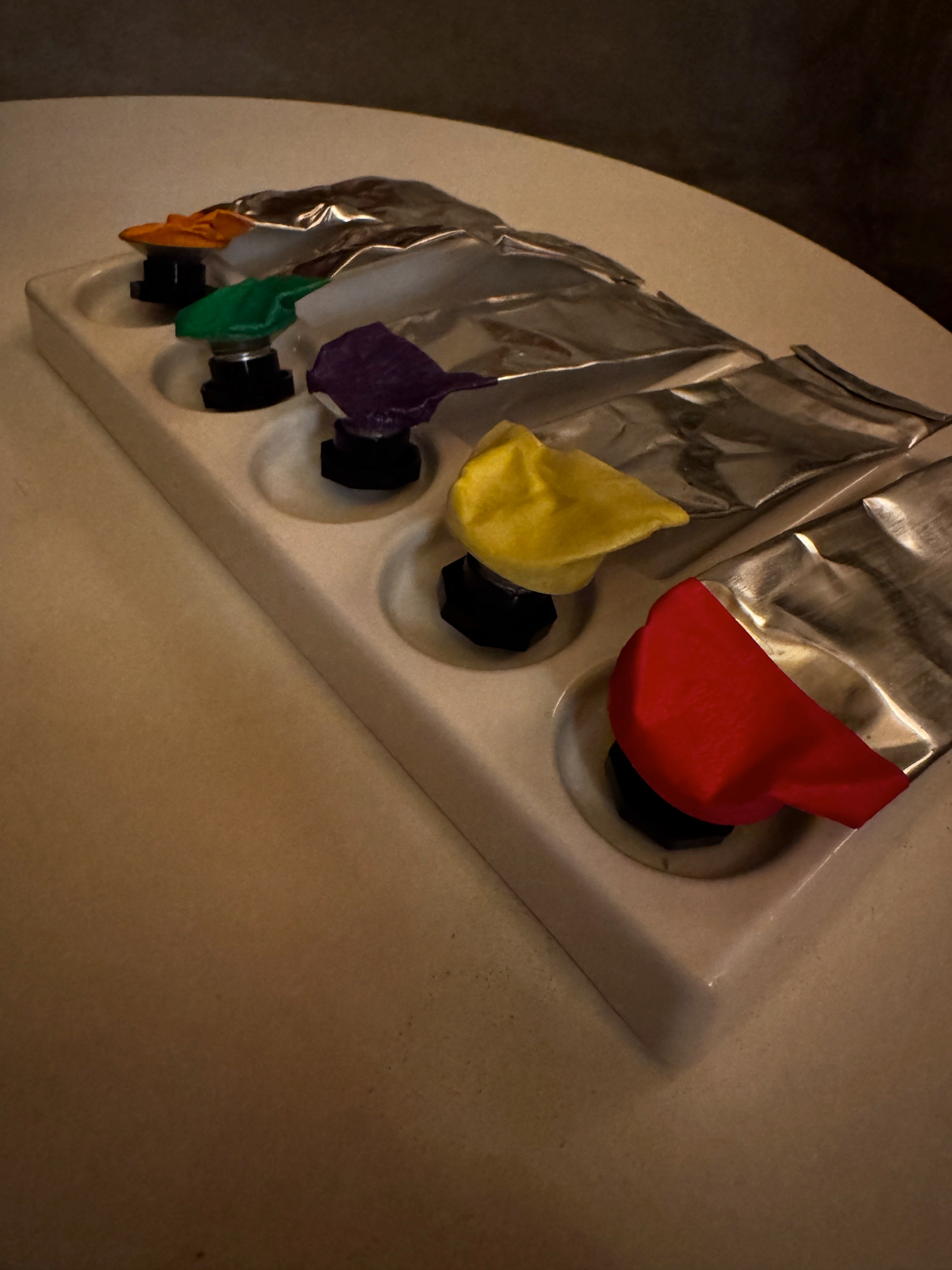 A row of small black droppers set into a white ceramic tray, each capped with a differently colored latex tip and partially covered with crinkled silver foil, suggesting precise, experimental cocktail preparation with a playful, theatrical presentation.