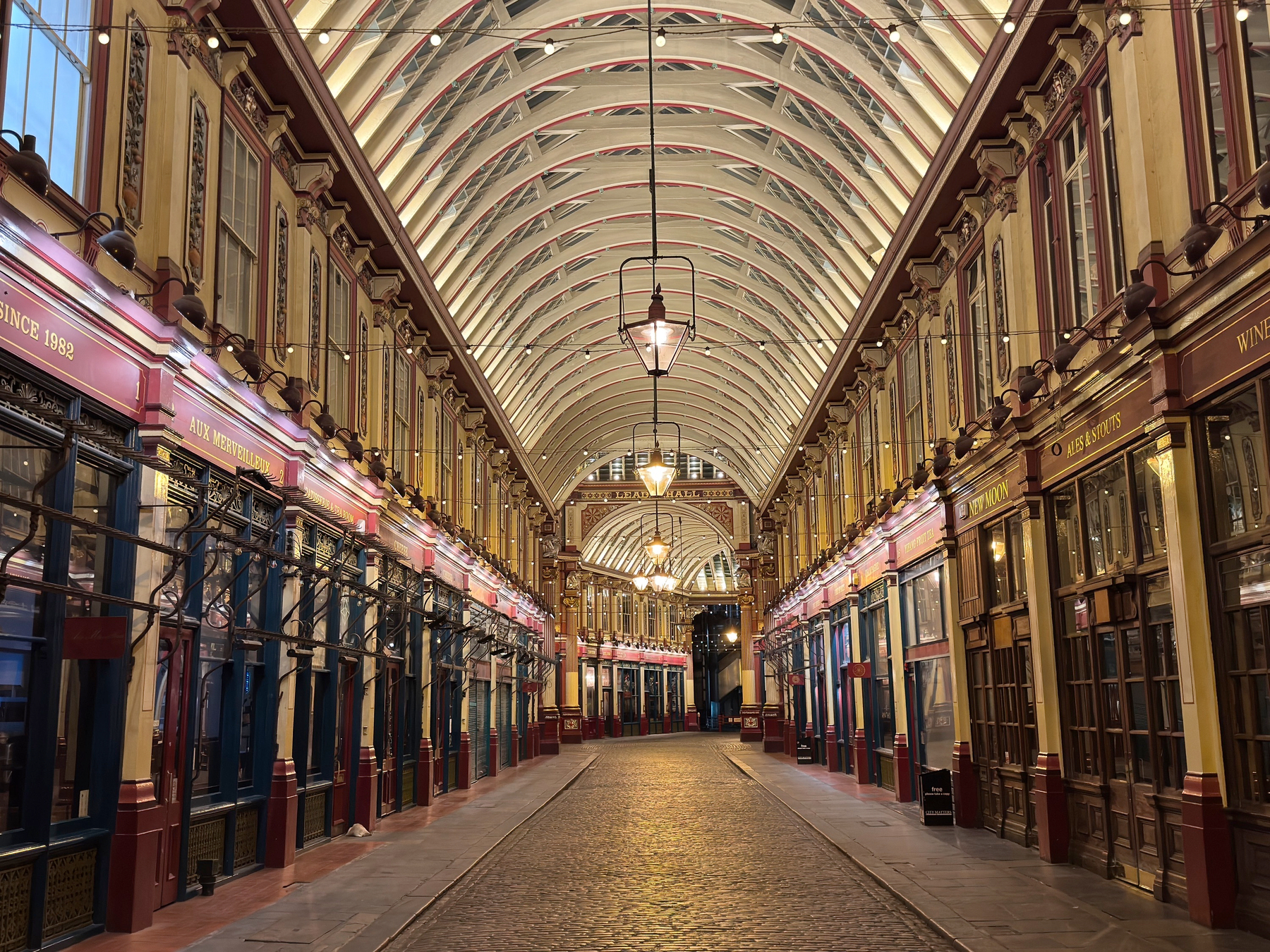 Symmetrical view down Leadenhall Market’s covered arcade, featuring ornate painted storefronts, a vaulted glass roof, hanging lanterns, and a quiet cobblestone walkway.