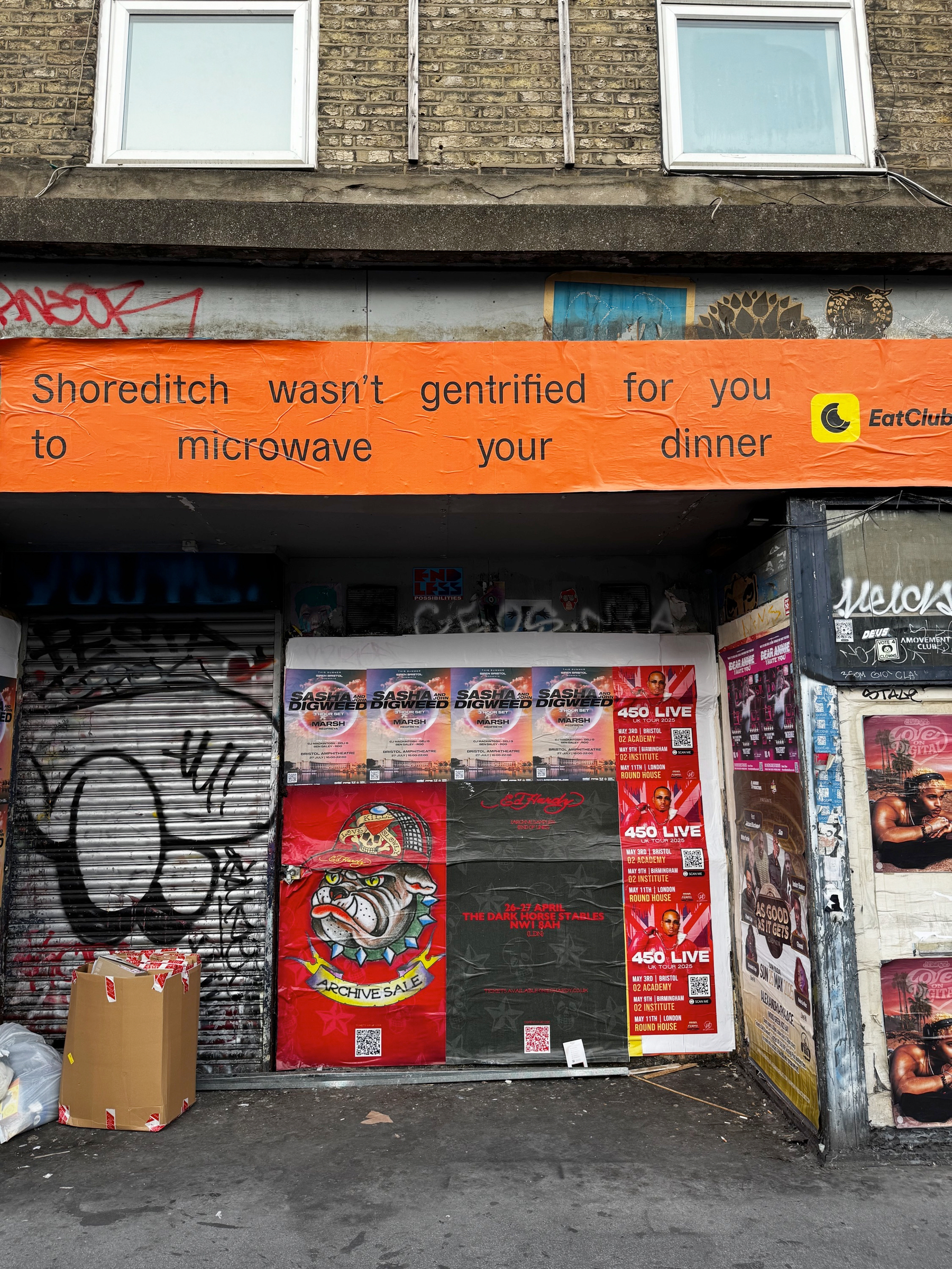 Street-level view of a shuttered storefront covered in posters and graffiti, topped by an orange banner reading “Shoreditch wasn’t gentrified for you to microwave your dinner,” with taped boxes and layered textures adding to the gritty scene.