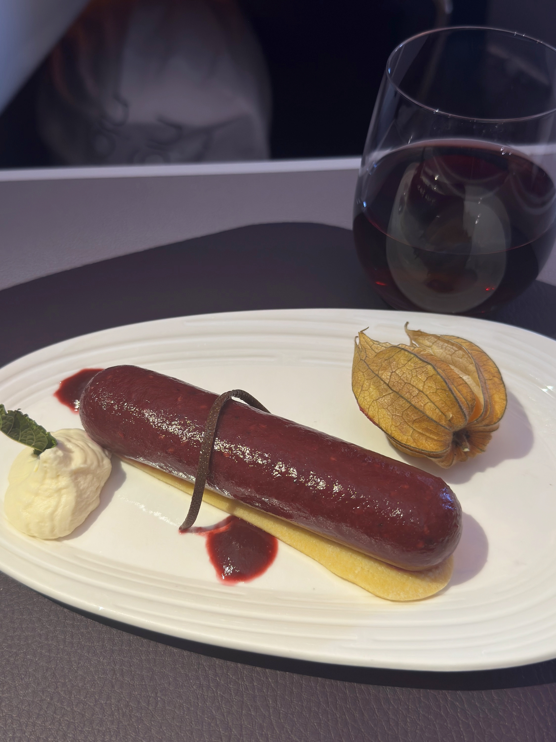 A refined dessert plate with a glossy dark berry parfait resting on sponge, paired with whipped cream, berry sauce, and a glass of red wine.
