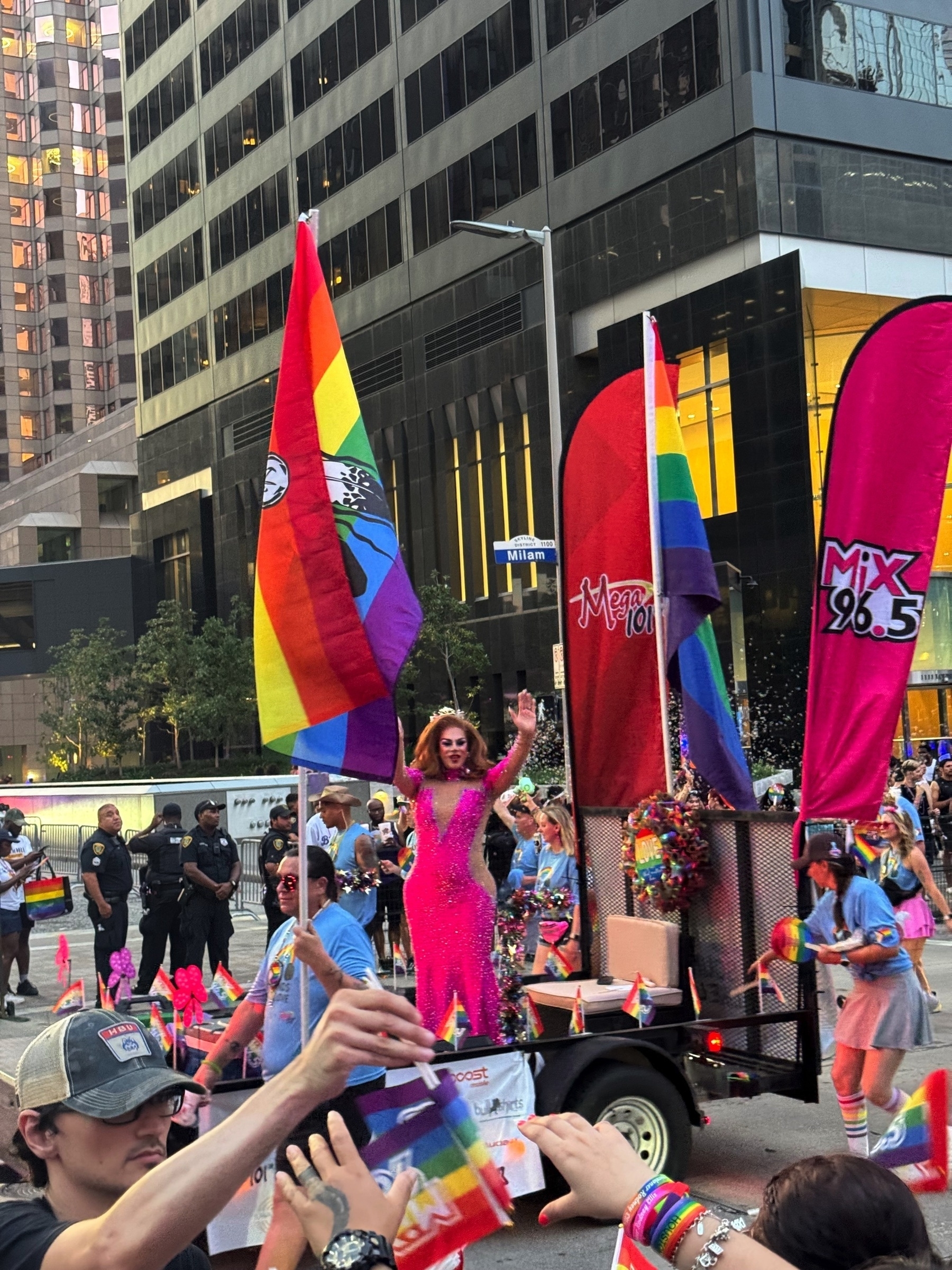 A drag performer in a sparkling hot pink gown waves from a parade float decorated with rainbow flags and glittering streamers. The float is branded with local radio stations Mix 96.5 and Mega 101, and is surrounded by participants in matching light blue shirts. Spectators reach out with rainbow flags and phones, capturing the performance and adding to the energetic, joyful vibe of the scene.