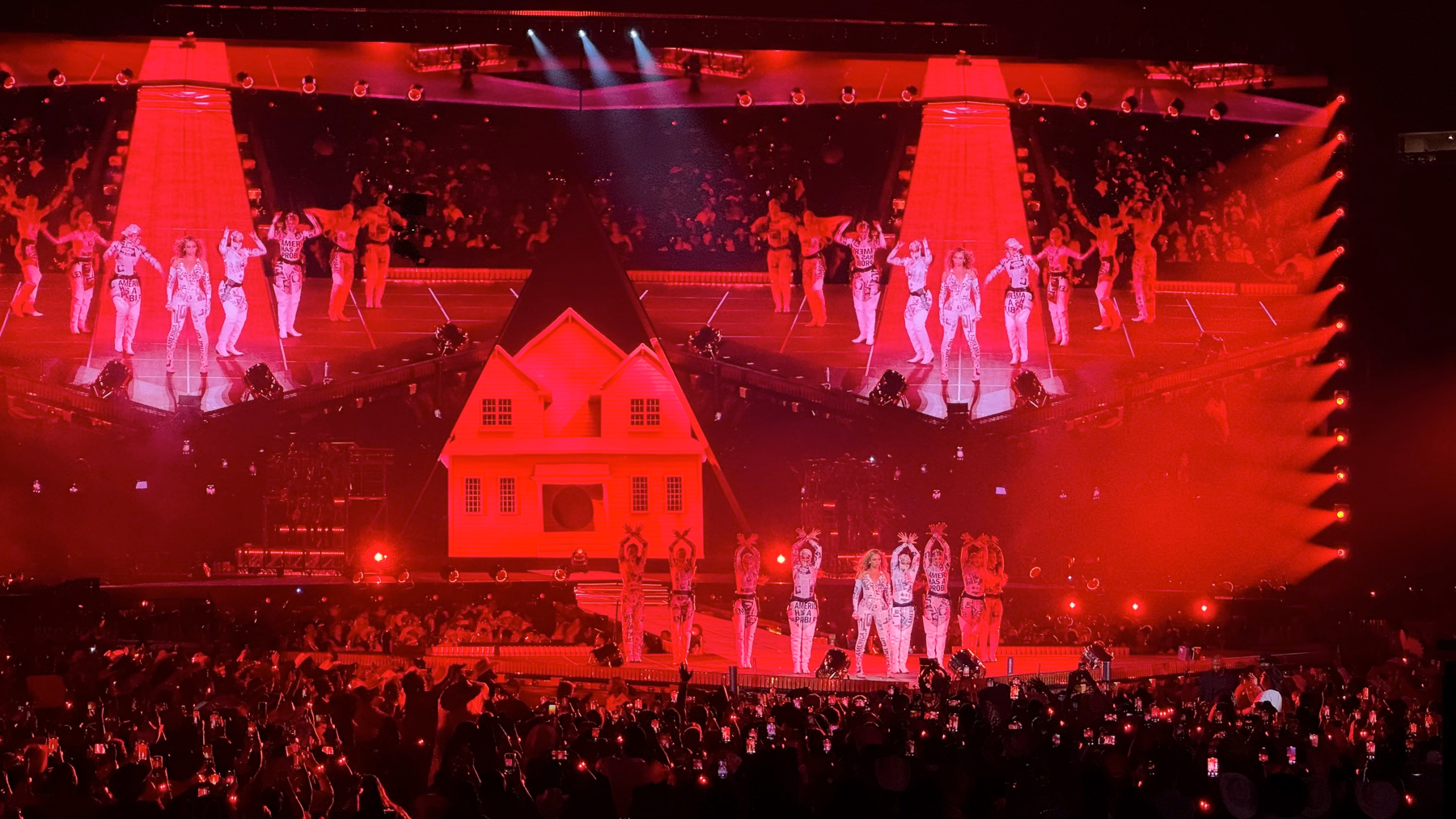 A full cast of dancers fills the stage beneath towering red lighting and a stylized house set, evoking Americana imagery. Beyoncé stands center as the performers strike synchronized poses, while the crowd below raises phones to capture the bold, graphic tableau.