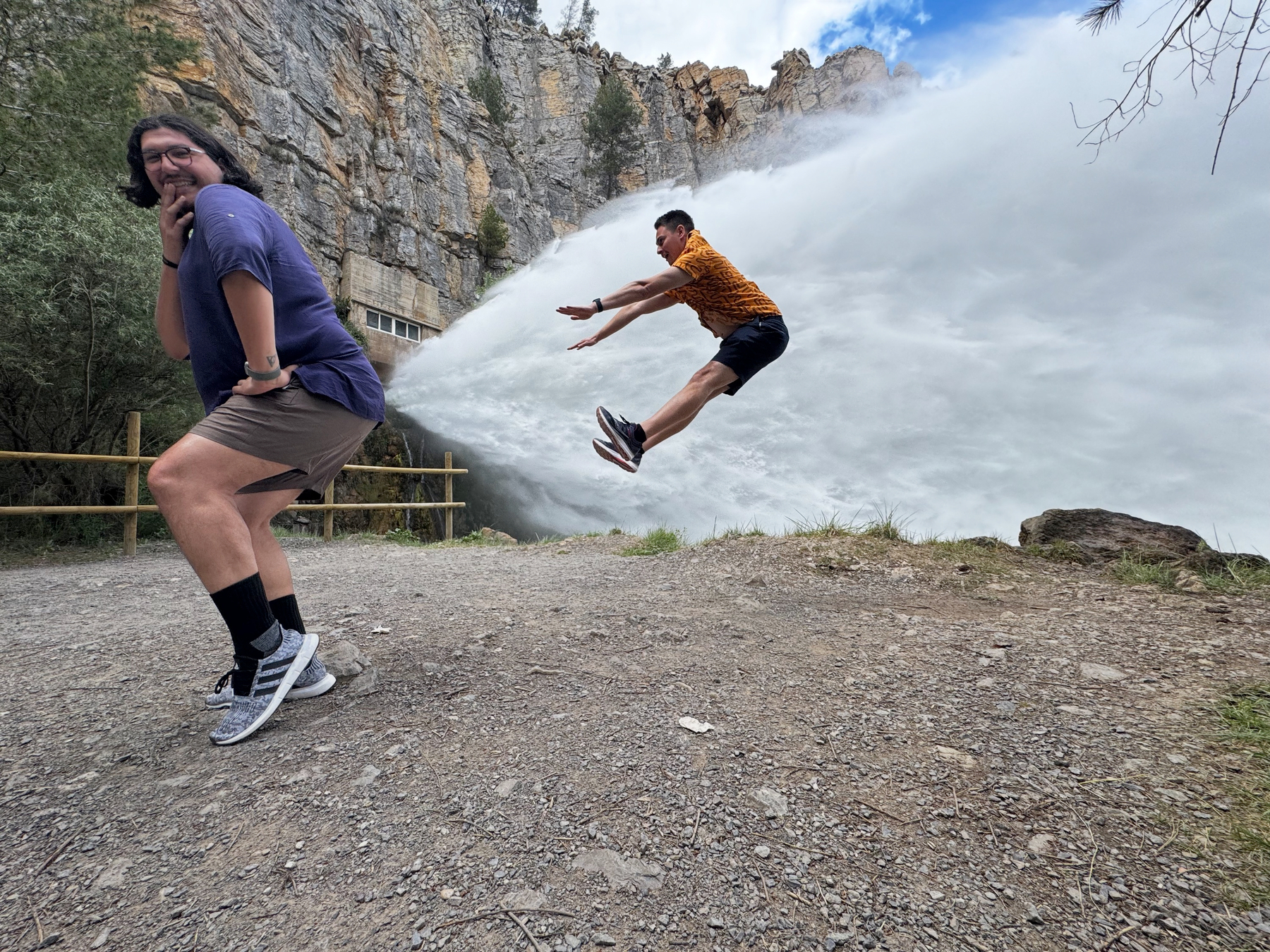 A playful action shot of Hugo and me near a roaring waterfall, Hugo crouching and laughing while I appear midair in a dramatic leap as water crashes down behind us.