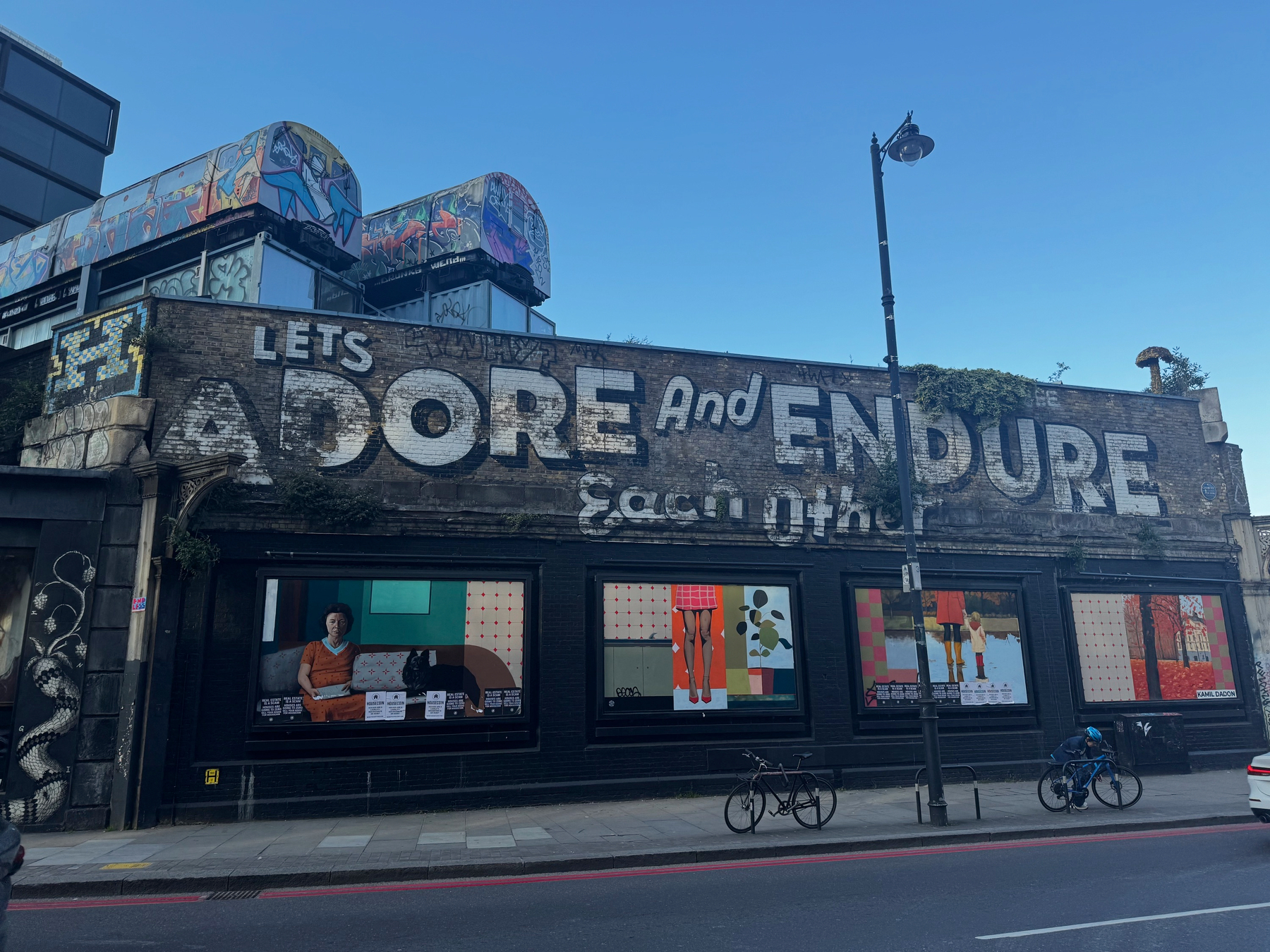 Wide view of a long brick building painted with the words “Let’s adore and endure,” large illustrated posters in the windows, bicycles parked outside, and traffic passing along the road.