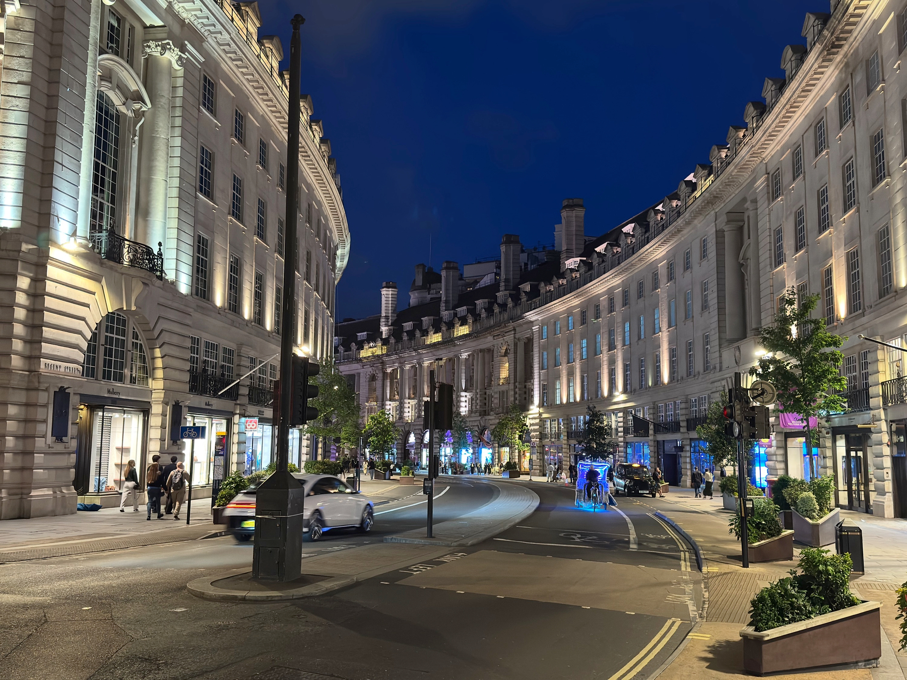 Curving sweep of Regent Street at night, with grand white stone buildings lit by storefronts and streetlights, light traffic, and pedestrians moving through the illuminated scene.