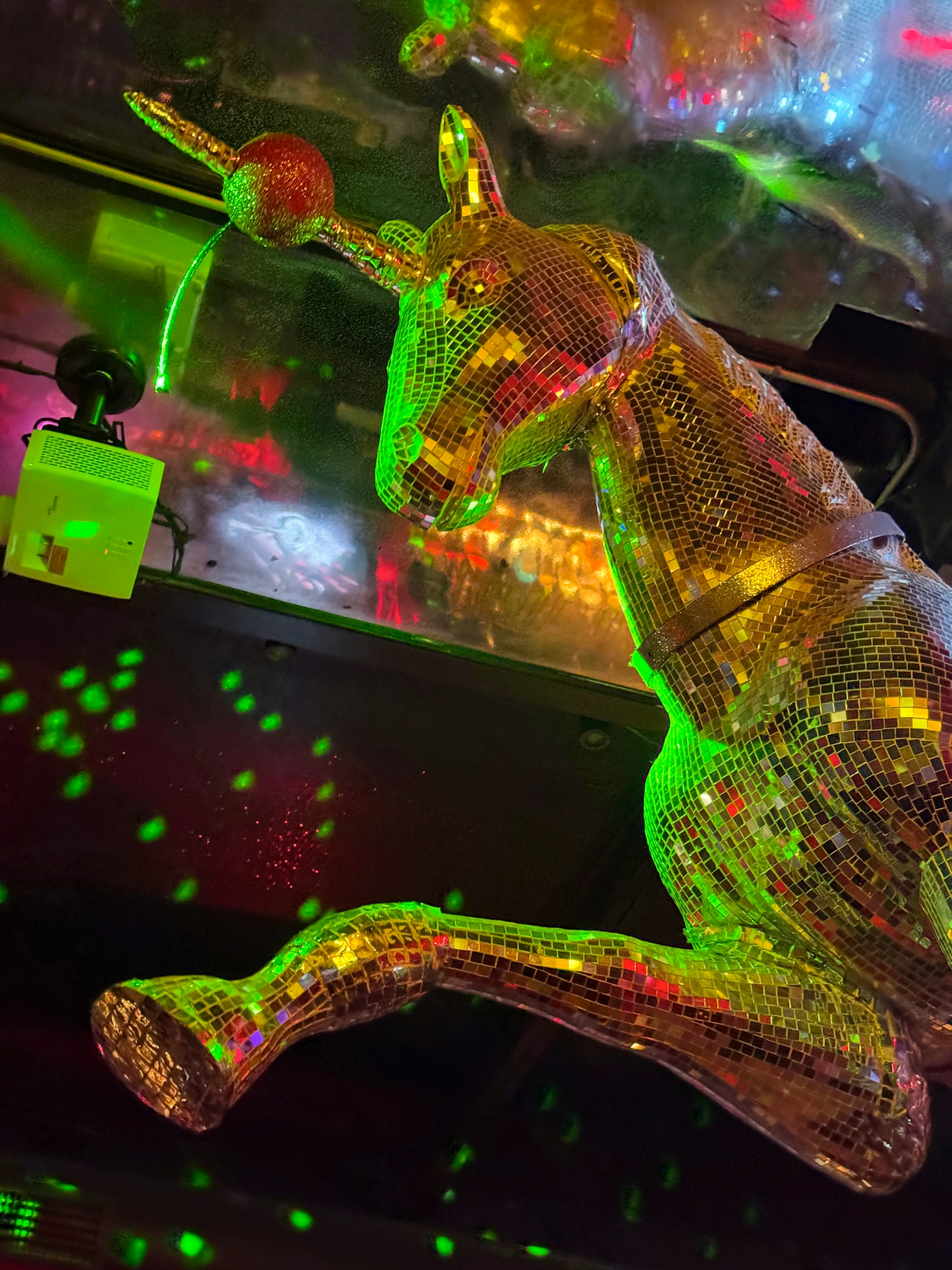 A mirrored unicorn sculpture suspended overhead, its mosaic surface reflecting green, red, and gold lights that scatter across the ceiling and surrounding space, creating a playful, dreamlike glow.
