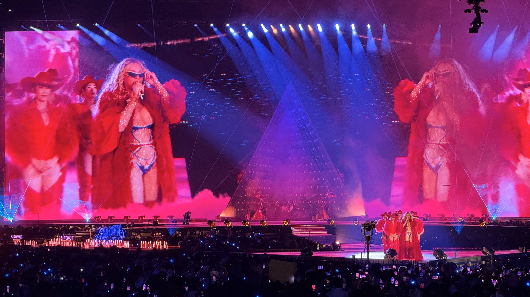 Beyoncé commands the stage in a dramatic red fur coat over a sparkling bodysuit, wearing dark sunglasses as dancers in coordinated red outfits flank her. Blue and purple stage lights contrast with the red costumes, and smoke effects add depth and glamour to the performance.