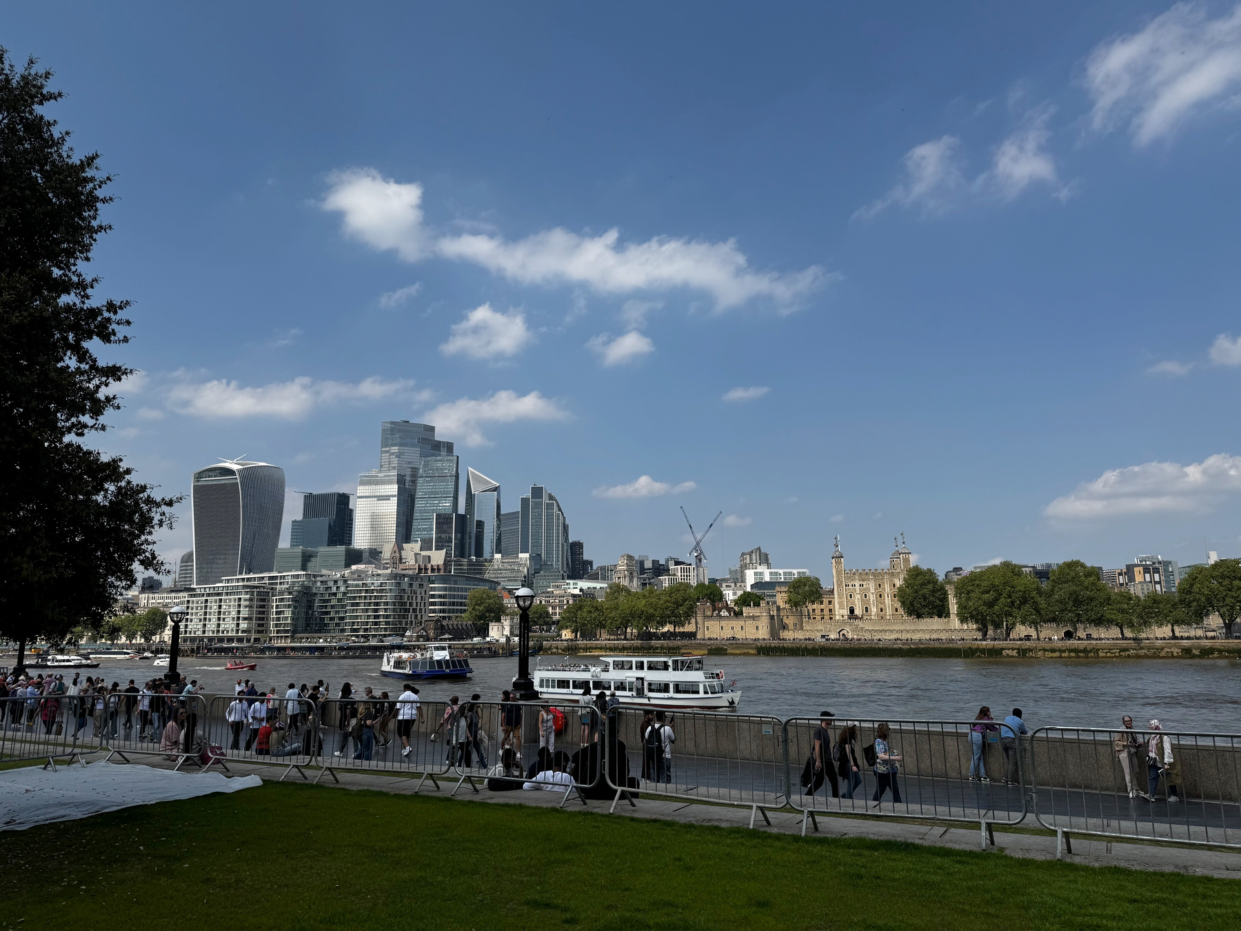 View across the River Thames showing modern glass skyscrapers on one side and the historic Tower of London on the other, with boats on the water and people lining the riverside path.