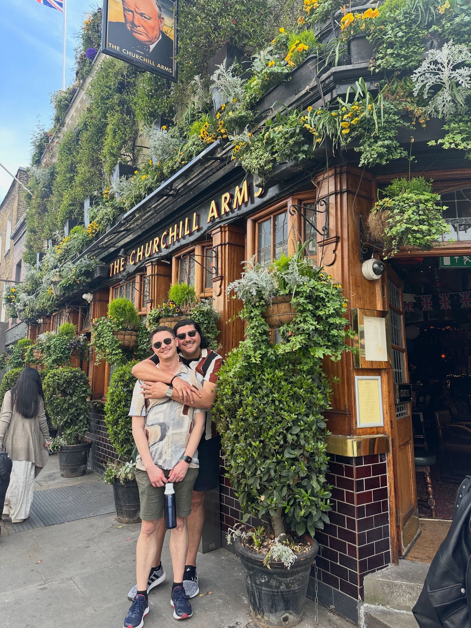 Hugo hugging me outside the pub entrance, standing beside tall potted plants and dark tiled walls, with menus by the door and greenery wrapping the façade.