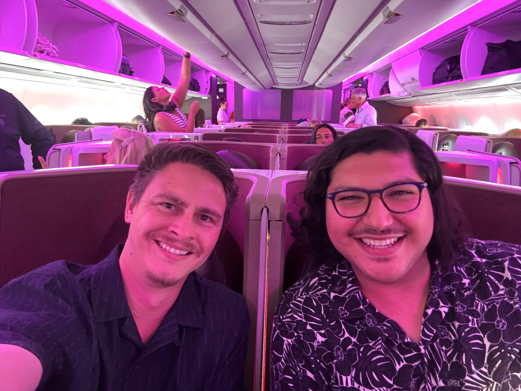 Hugo and I smiling while seated side by side in business class under soft purple cabin lighting, with other passengers settling in behind them.