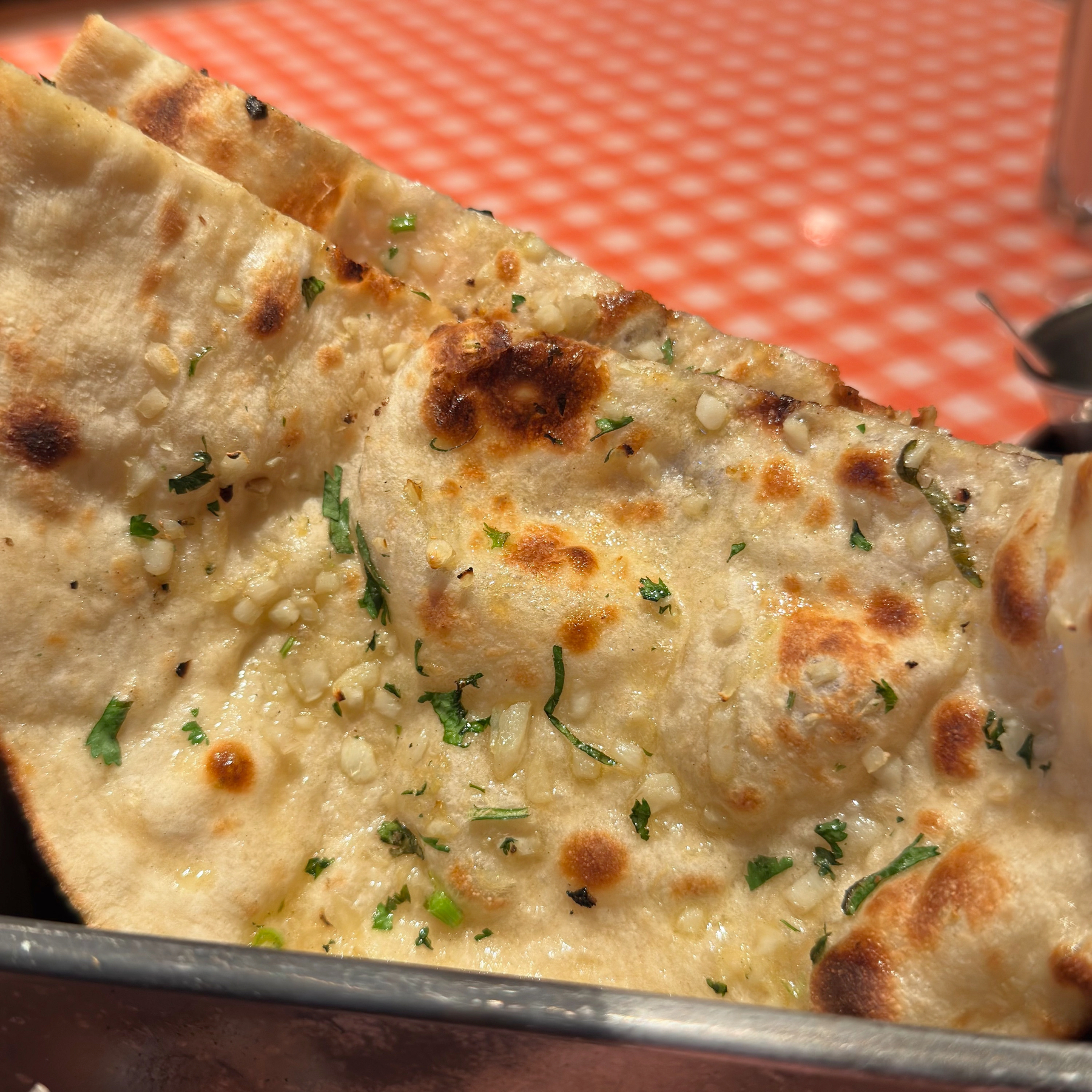 Soft, golden naan bread with blistered bubbles and crisp char marks rests in a metal tin, brushed with butter and scattered with chopped garlic and herbs. The surface looks pillowy and glossy, suggesting warmth, chew, and indulgent flavor.