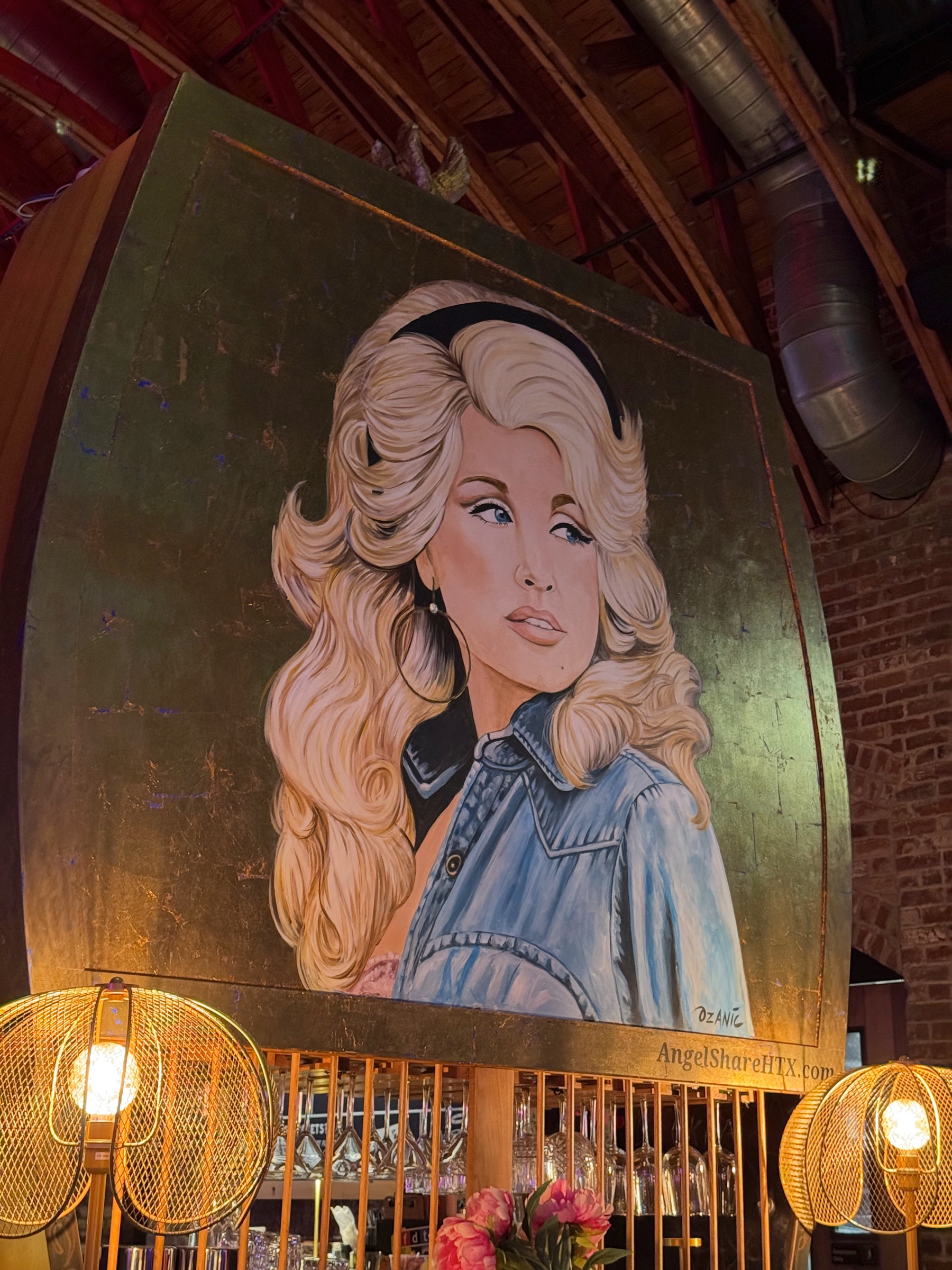 A large painted portrait of Dolly Parton with voluminous hair and hoop earrings, wearing a denim jacket and gazing to the side, mounted above a warmly lit bar with hanging lamps and exposed brick.
