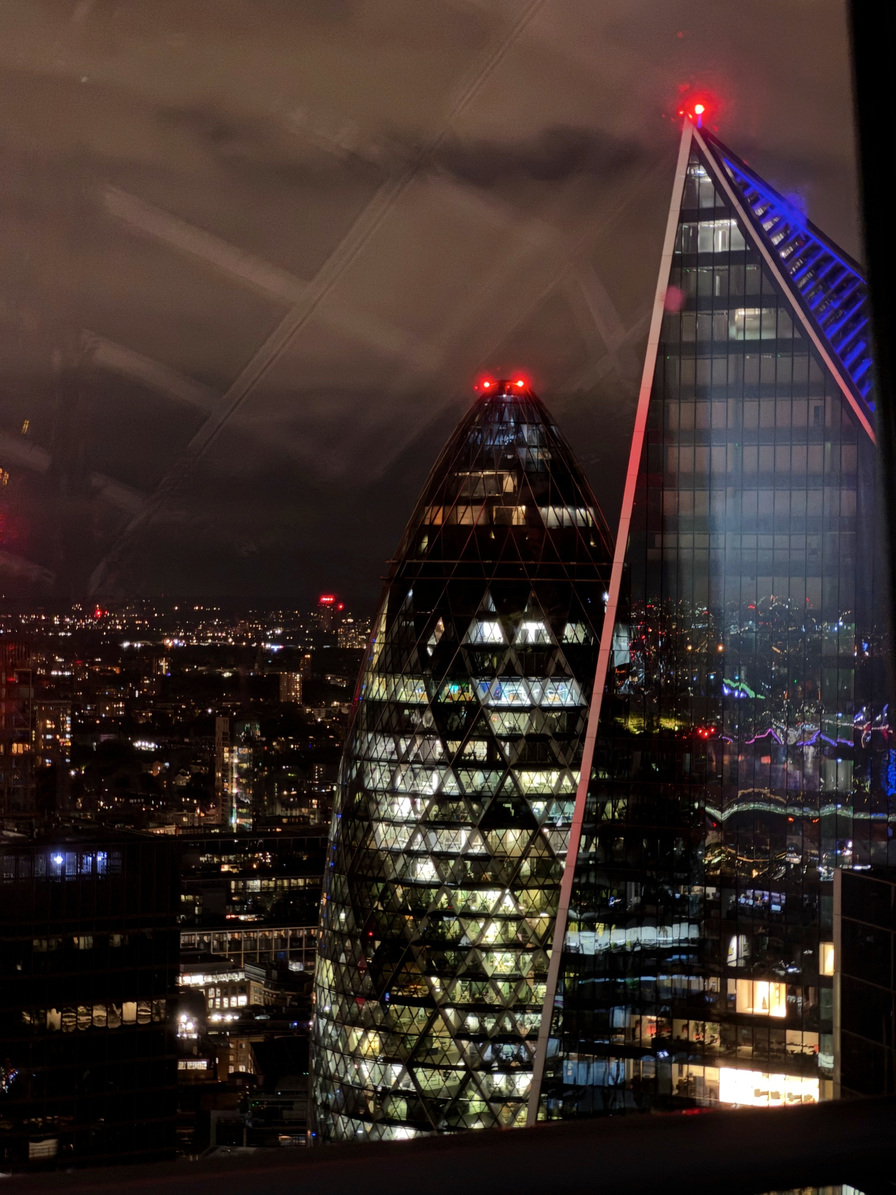 Night view over the City of London featuring the illuminated Gherkin and a sharp, angular glass tower, their office lights forming geometric patterns against a cloudy sky.