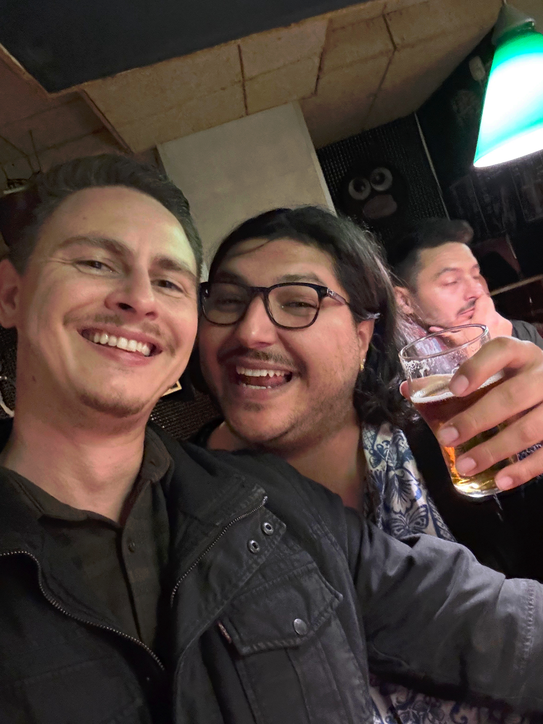 Hugo and I smiling closely together inside a busy bar, one holding a glass of amber beer, with warm indoor lighting and other patrons crowded in the background.