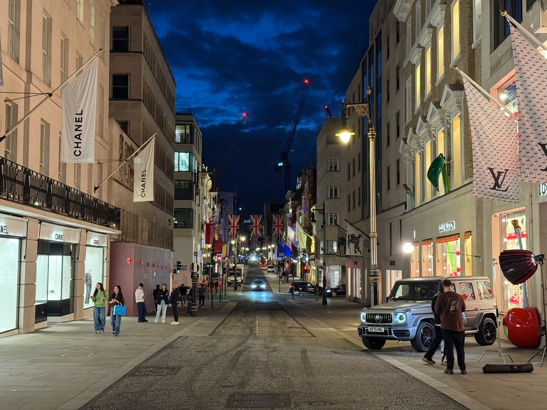 Evening view down Bond Street with luxury storefronts, Chanel flags, Union Jack banners, passing cars, and pedestrians under a deep blue sky.