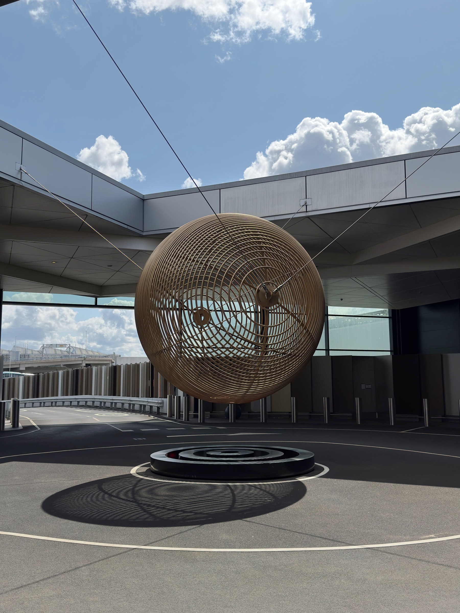 A large woven metal sphere sculpture suspended by cables in an open-air terminal courtyard, casting intricate shadows on the ground beneath a bright blue sky with billowing white clouds.